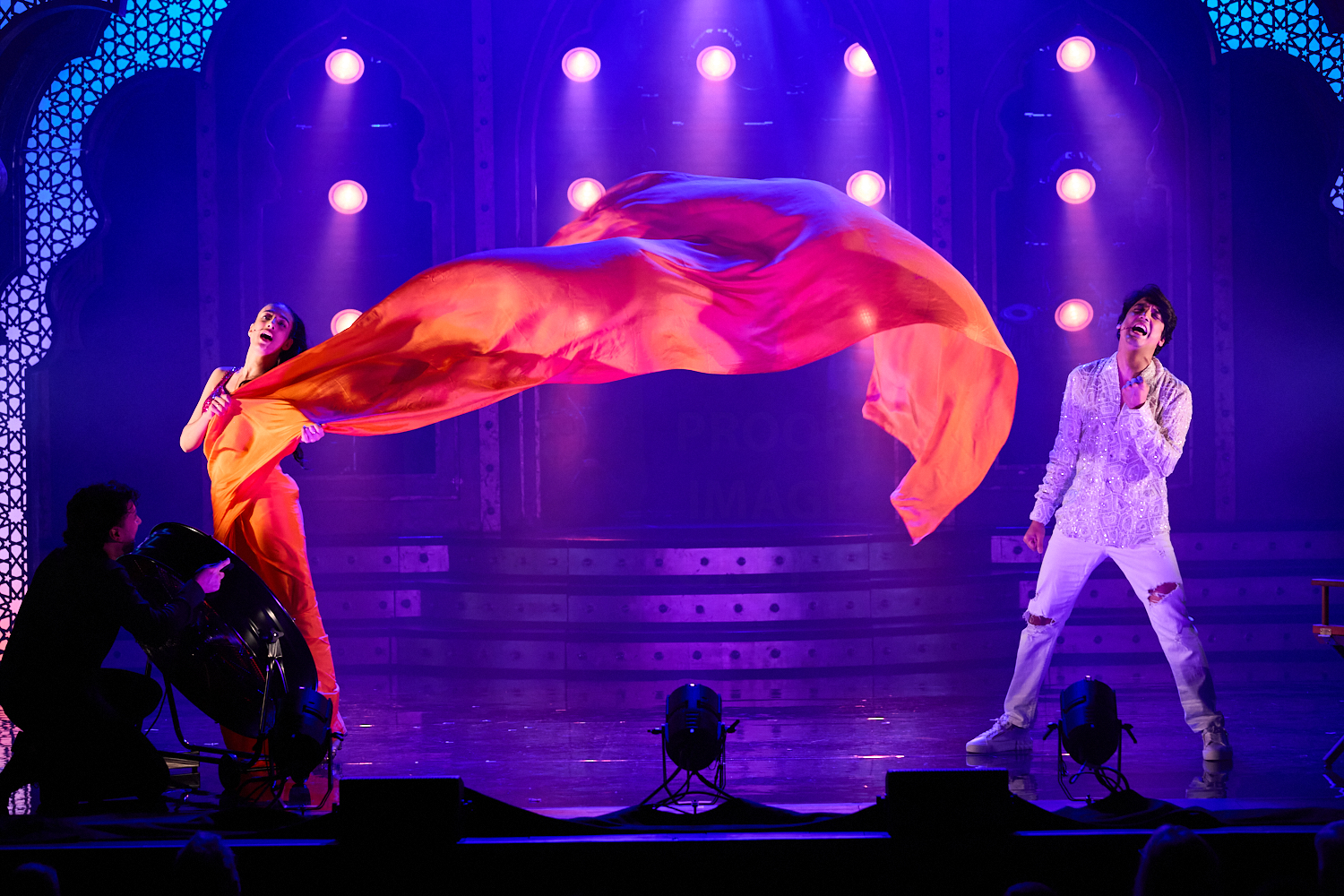 A vibrant performance with a woman in an orange dress holding a flowing fabric on stage. A man in a patterned shirt sings passionately.