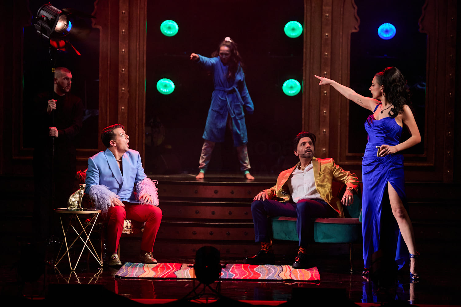 A theatrical scene shows two men seated, one in a light blue jacket with pink feathers, the other in a gold jacket. A woman in a blue gown points at them emphatically while another woman, in a blue robe, stands behind on a staircase pointing as well. The atmosphere is dramatic and intense, with colorful lights in the background.