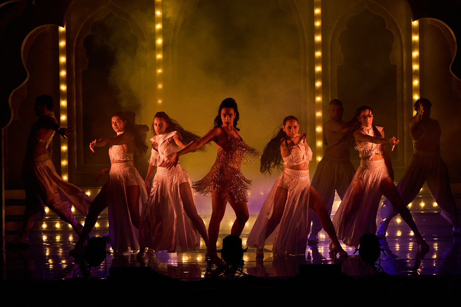 A group of dancers performs on stage, with a central figure in a shimmering gold outfit. The stage is lit dramatically, with warm, glowing arches and an intense, vibrant energy.