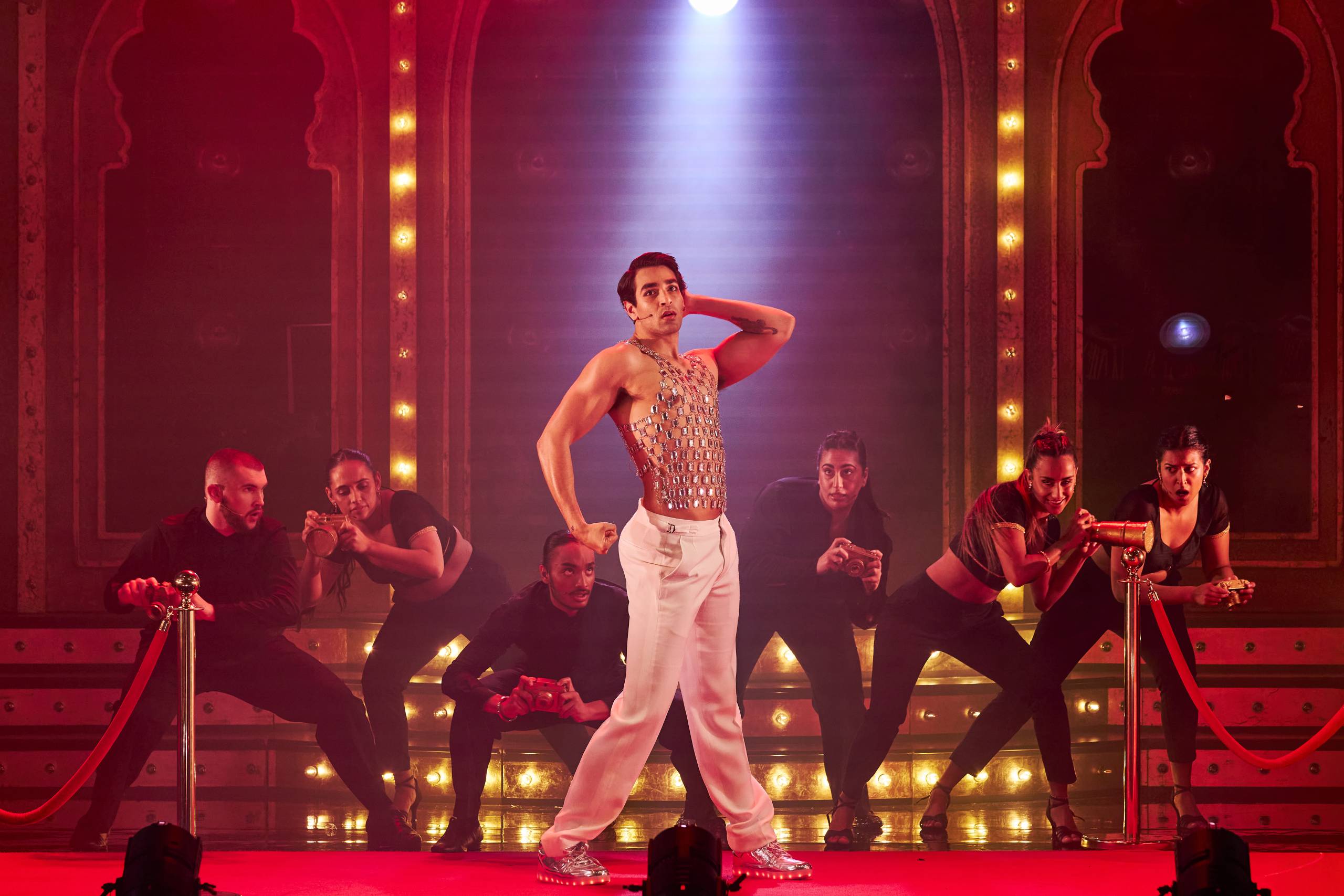 A performer in a sparkly top and white pants poses confidently on a stage, surrounded by dancers. The background features bright lights and red curtains, creating a vibrant, theatrical atmosphere.