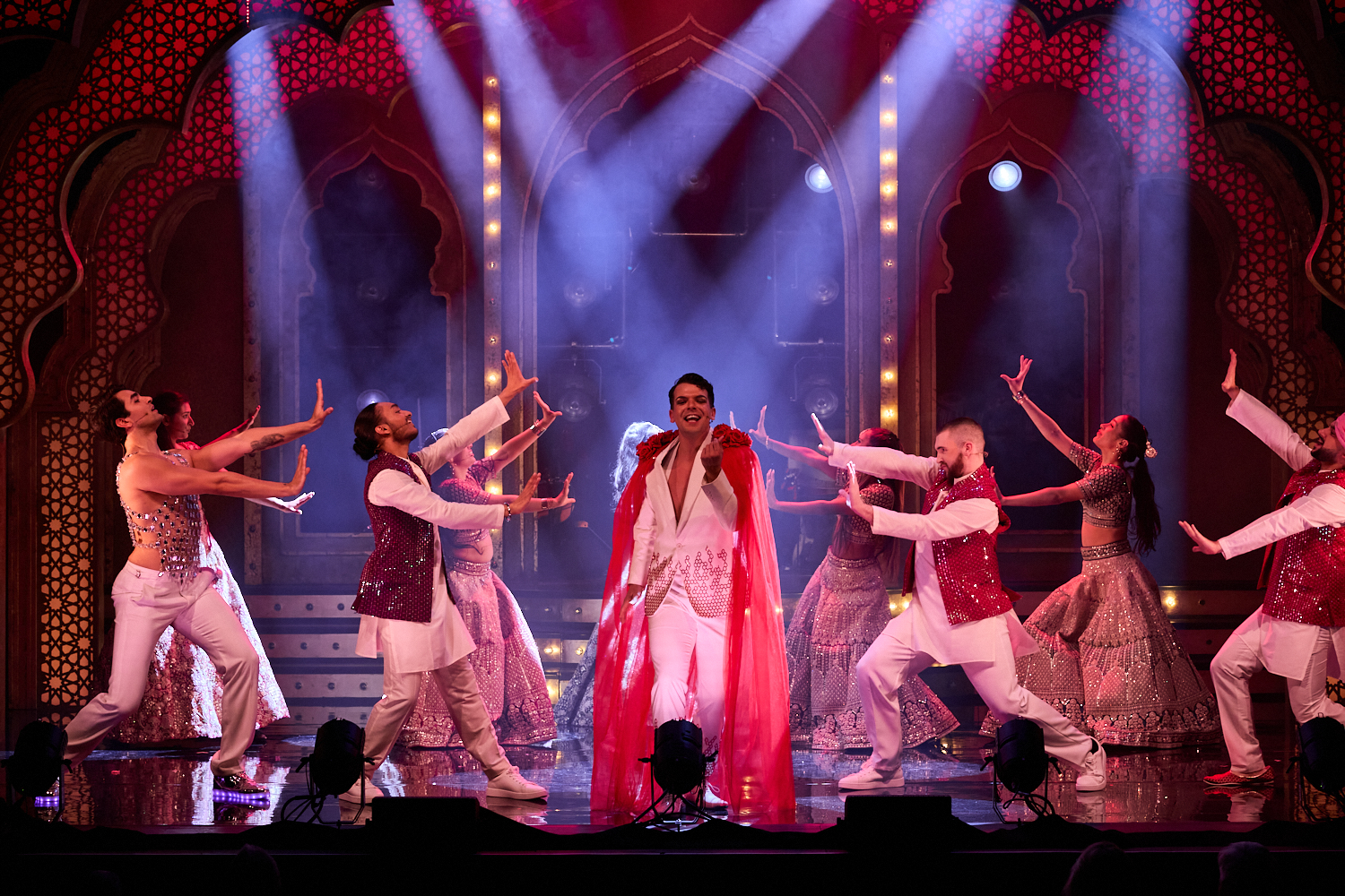 Performers in vibrant costumes dance energetically on a stage with ornate arches. Dramatic lighting and joyful expressions highlight a festive atmosphere.