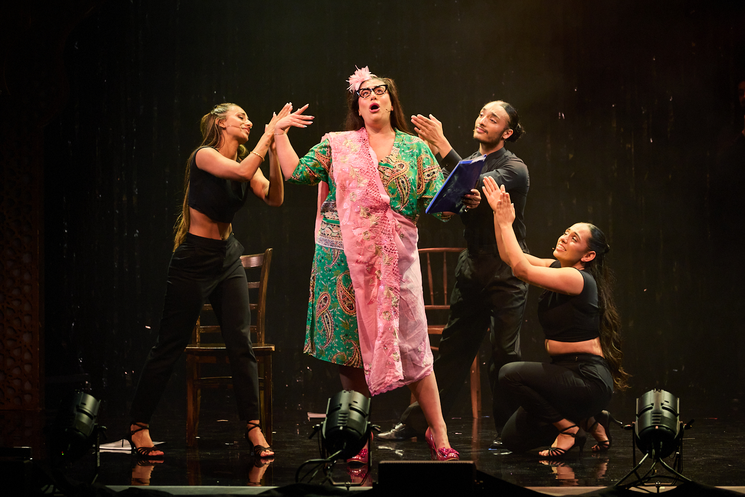 A person in a colorful robe and glasses performs dramatically on stage with three backup dancers in black outfits. The scene is lively and theatrical.