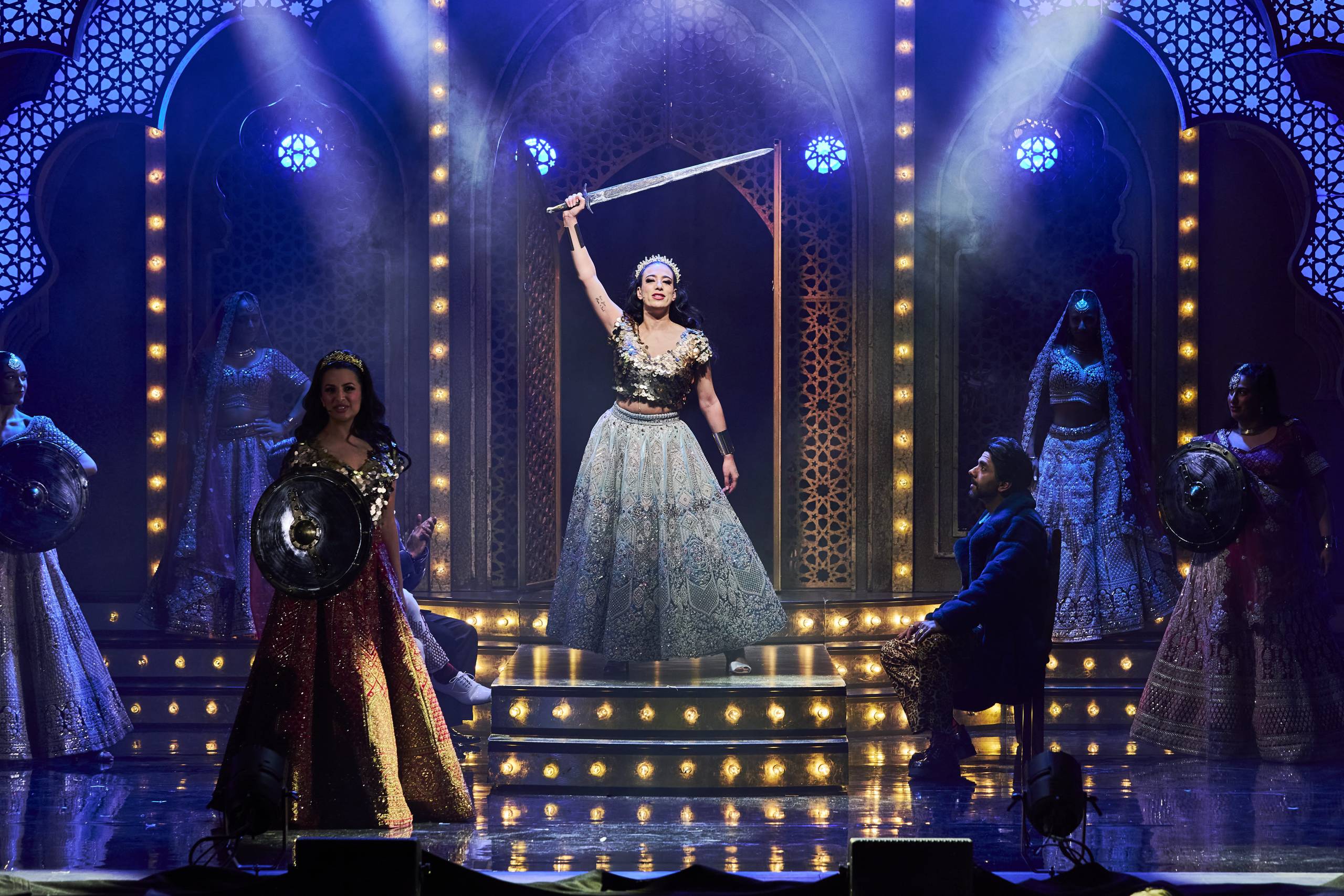 A woman in a glittering gown holds a sword aloft on a brightly lit stage, surrounded by performers in regal costumes, creating a dramatic scene.