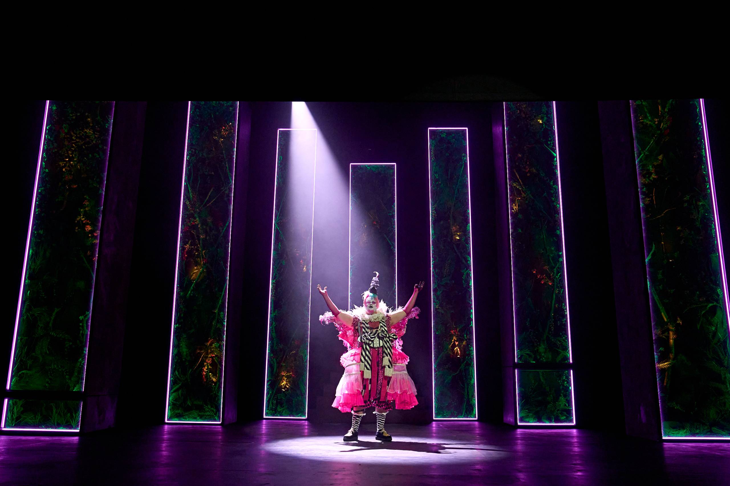 Puck in a vibrant, theatrical costume stands under a spotlight on a dark stage. Neon-lit panels frame the scene,