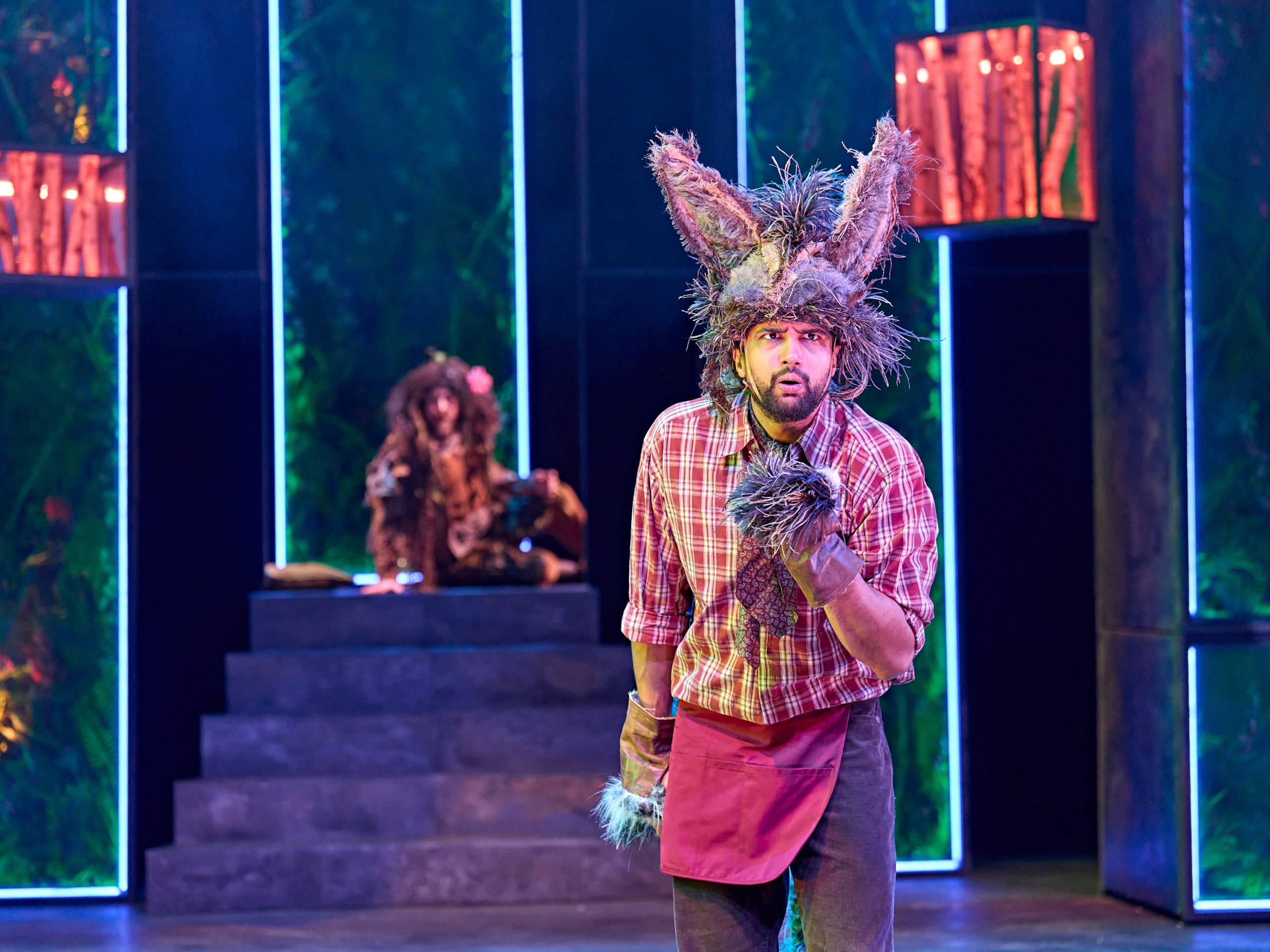 Bottom wearing a donkey headpiece and gloves, stands on stage with a surprised expression. Oberon sits in the background.