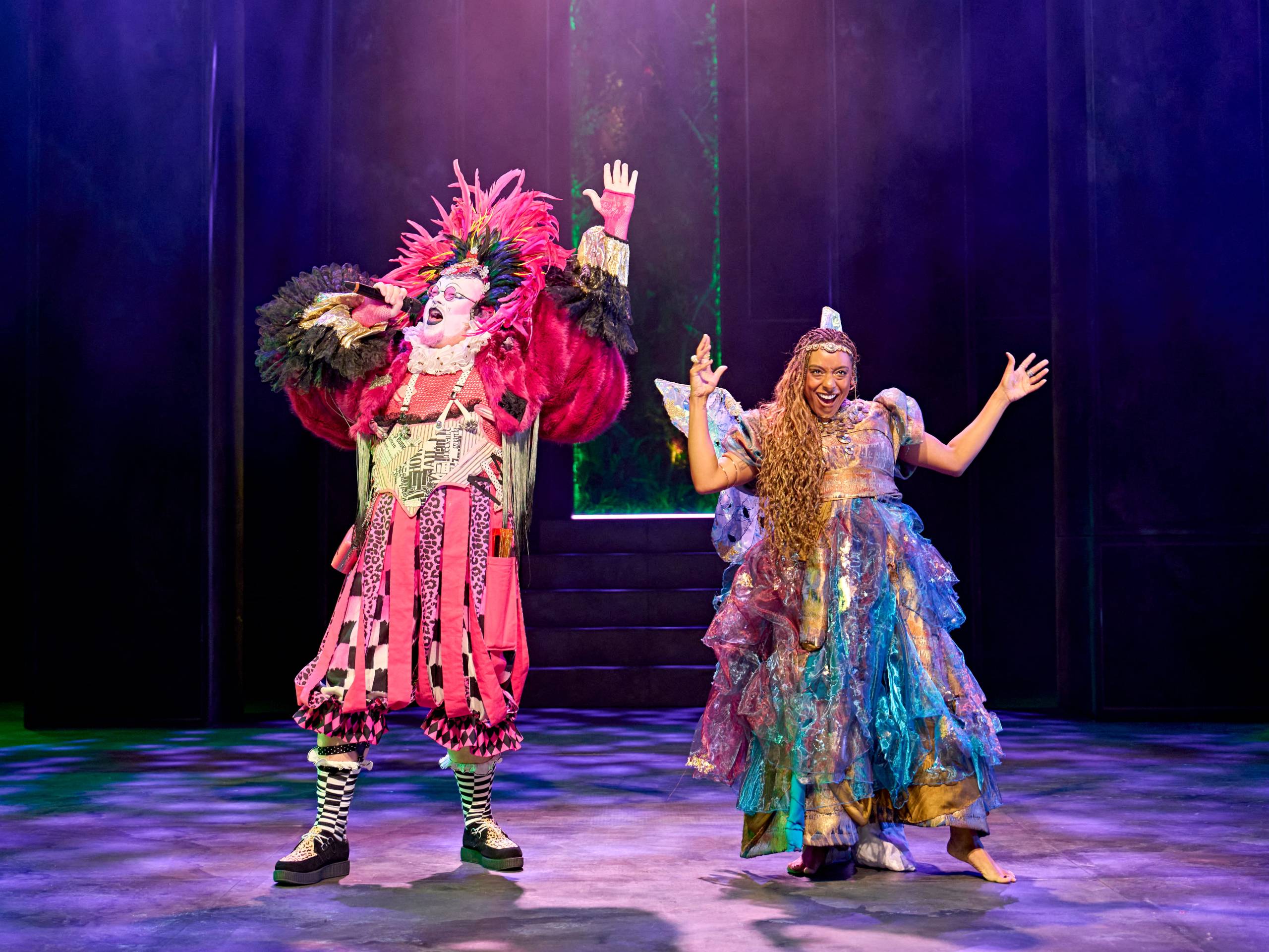 Two performers on stage wearing colourful extravagant costumes, Puck with a large headdress and Titania in a shimmering, layered dress.