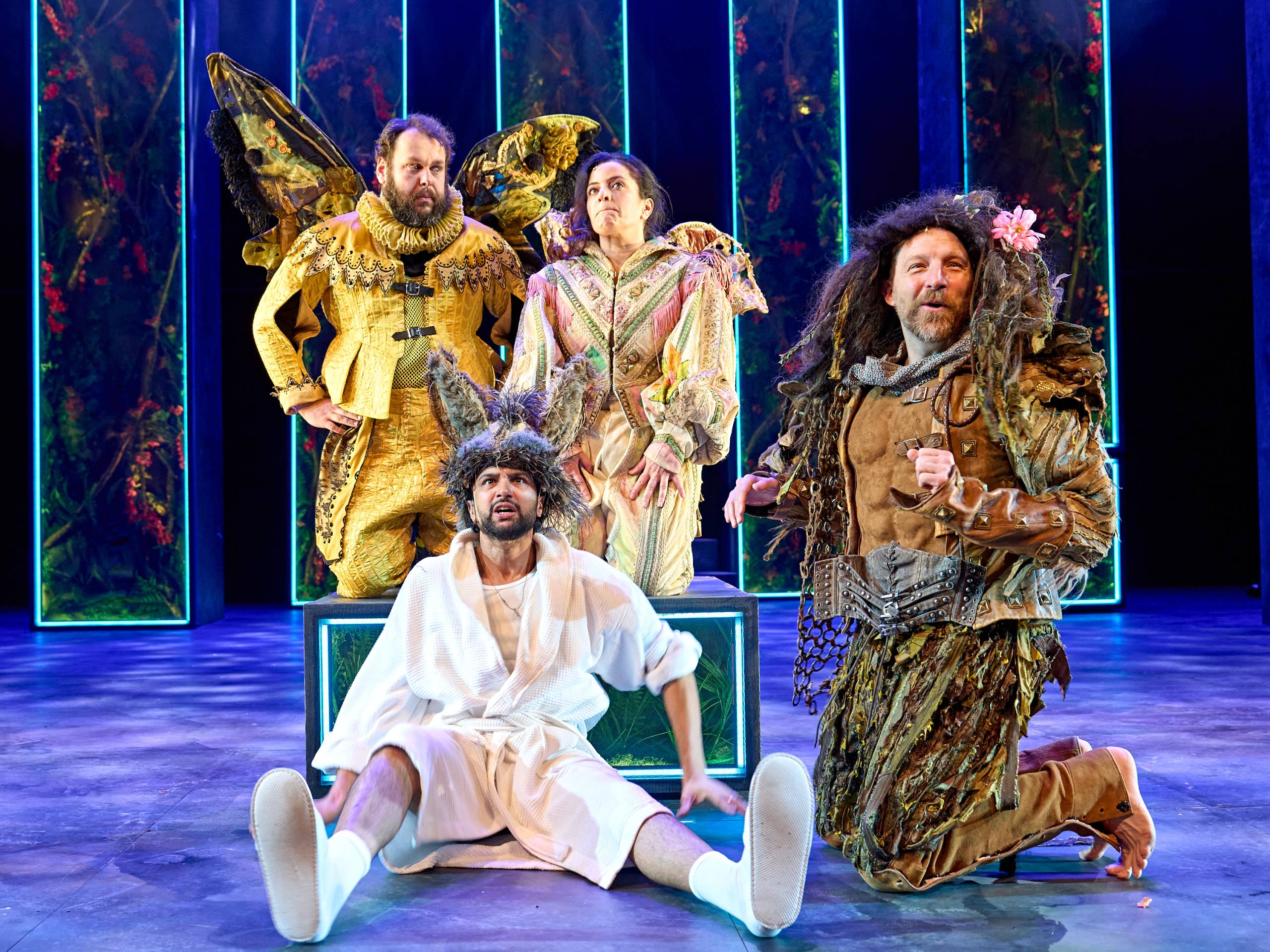 Bottom in a white robe with donkey ears sits in the foreground looking startled, while two colourful fairies and Oberon kneel and gesture expressively.
