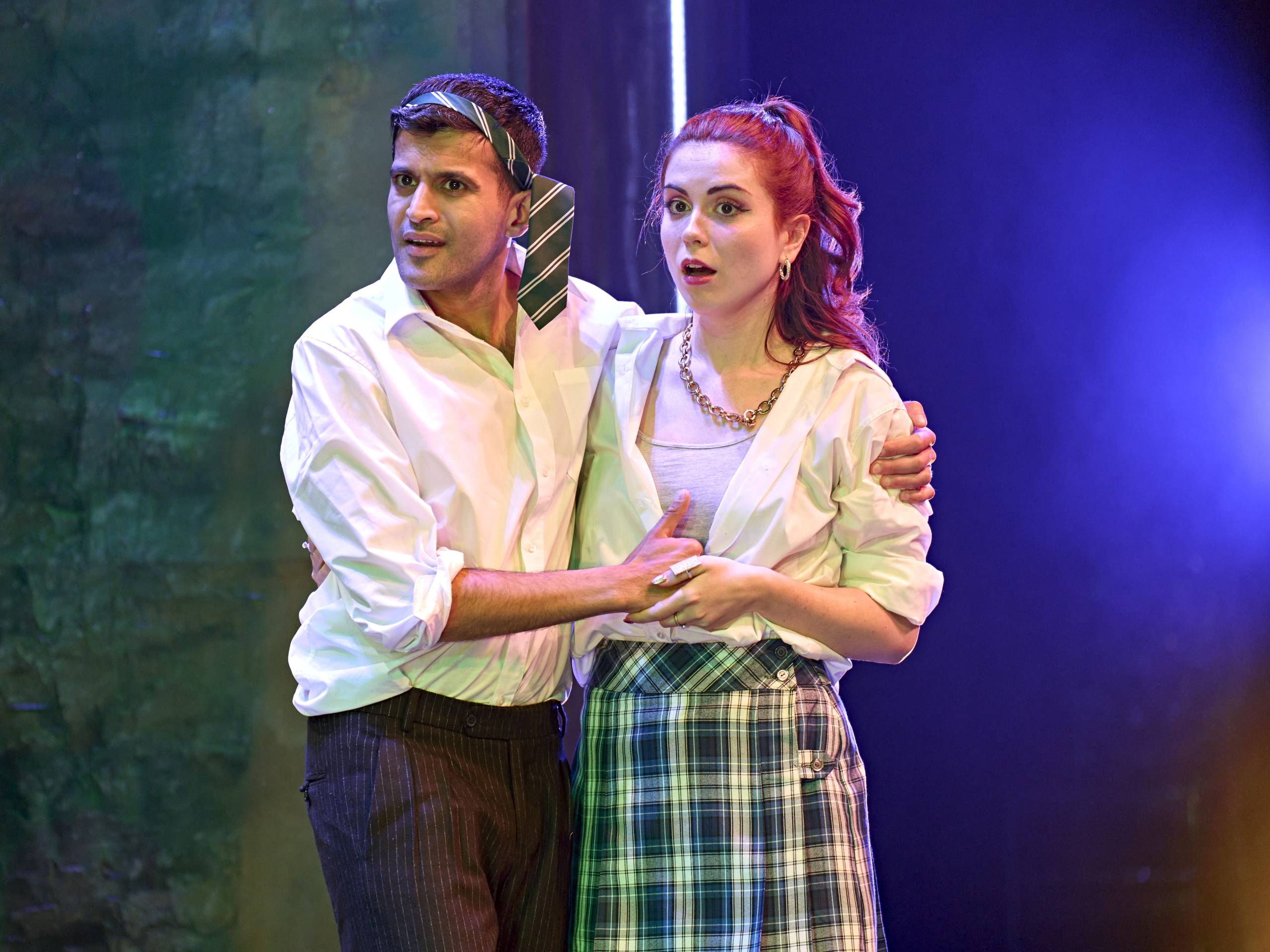 Demetrius and Helena stand close together. He wears a loose tie around his head and a shirt, and she wears a plaid skirt, open shirt and a necklace, both appearing surprised.