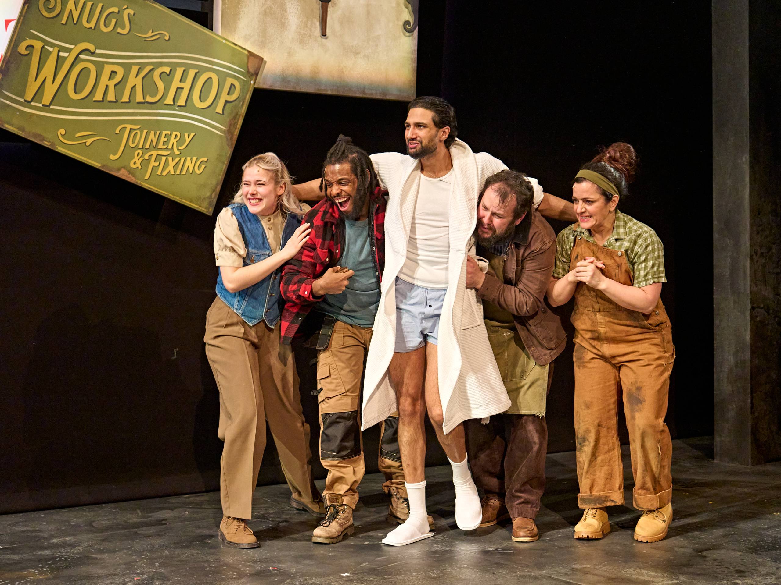 Five actors stand closely on stage, smiling and supporting each other under a 