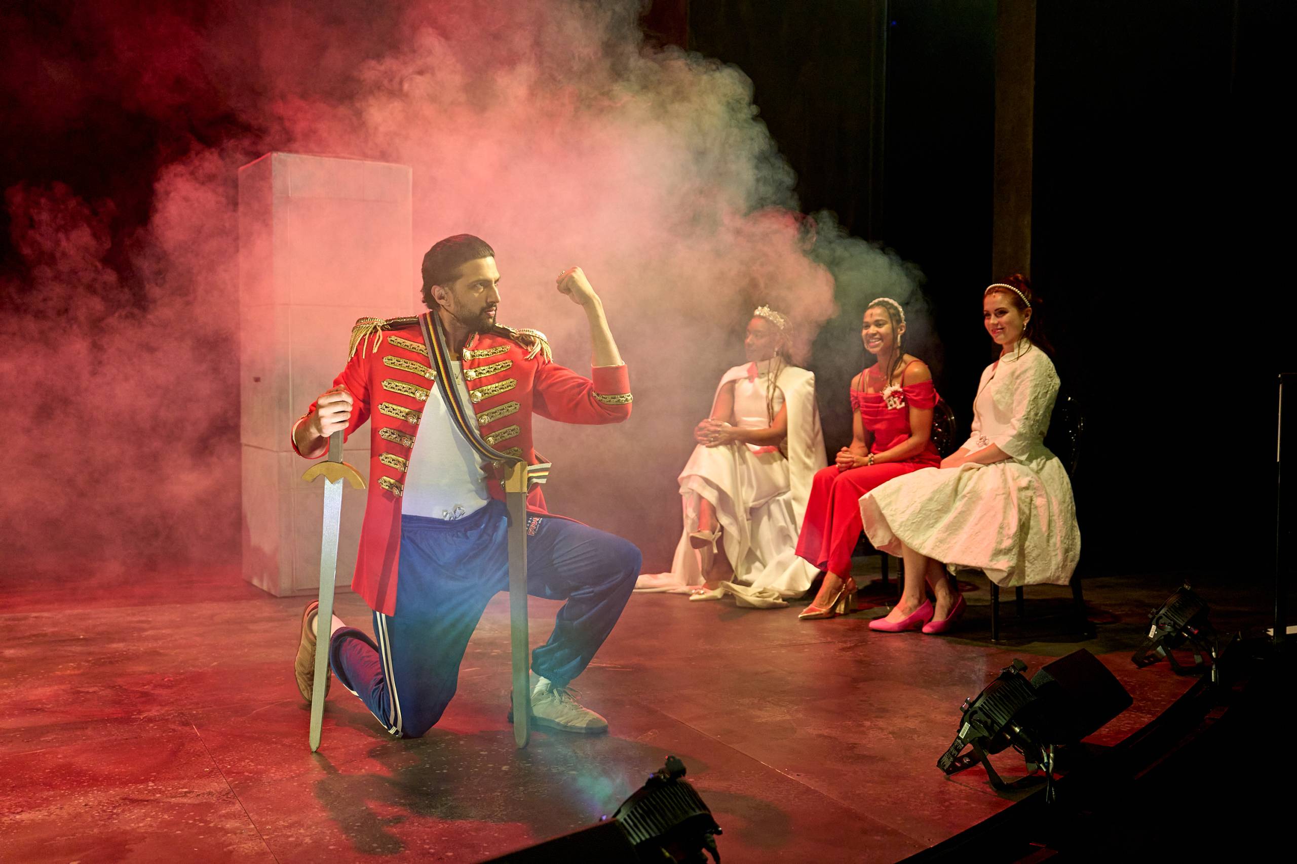 A performer in a vibrant red costume strikes a pose amid colourful stage smoke. Three women in elegant dresses watch.