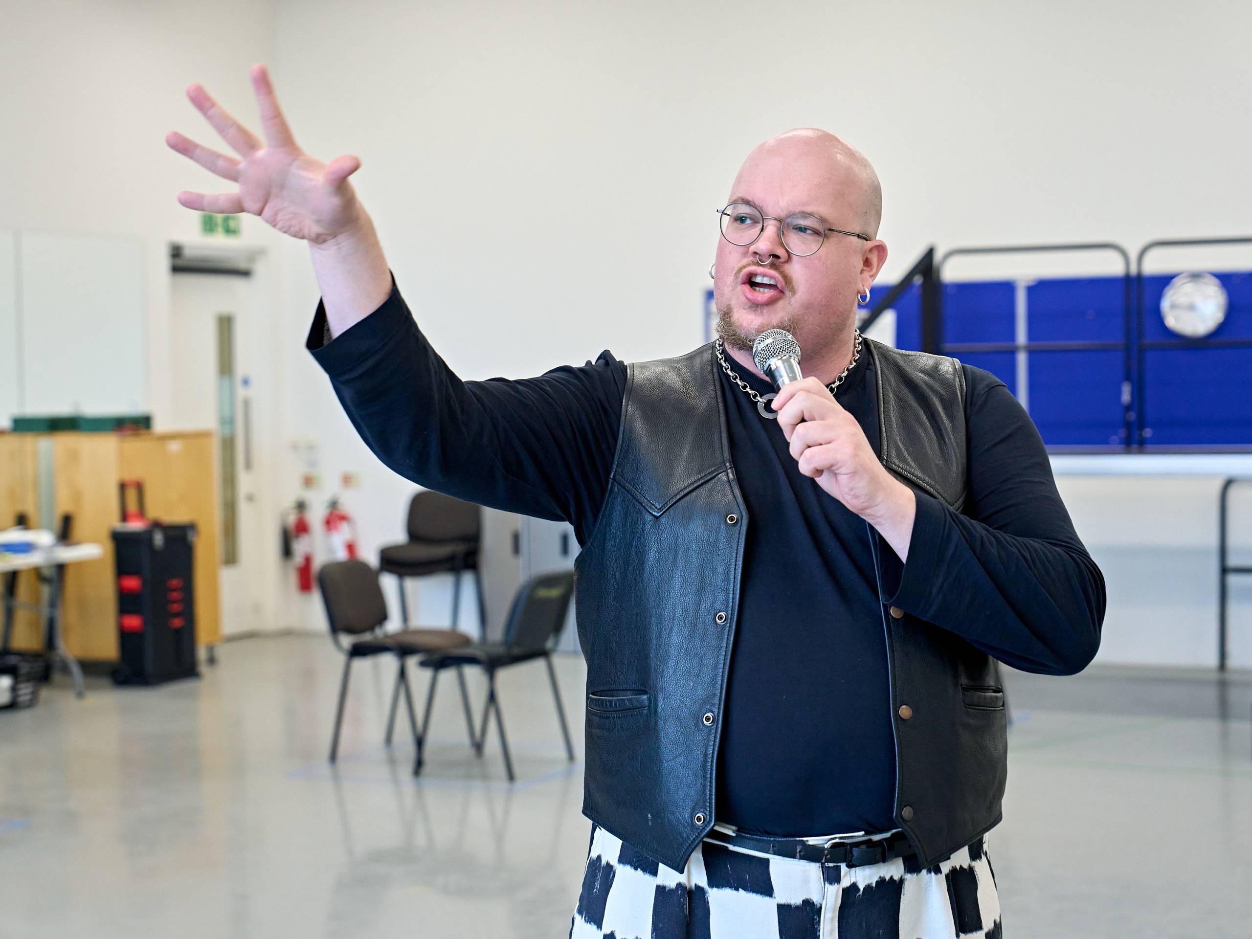 A person with glasses and a goatee is singing into a microphone, wearing a black leather vest and a black-and-white checked outfit.