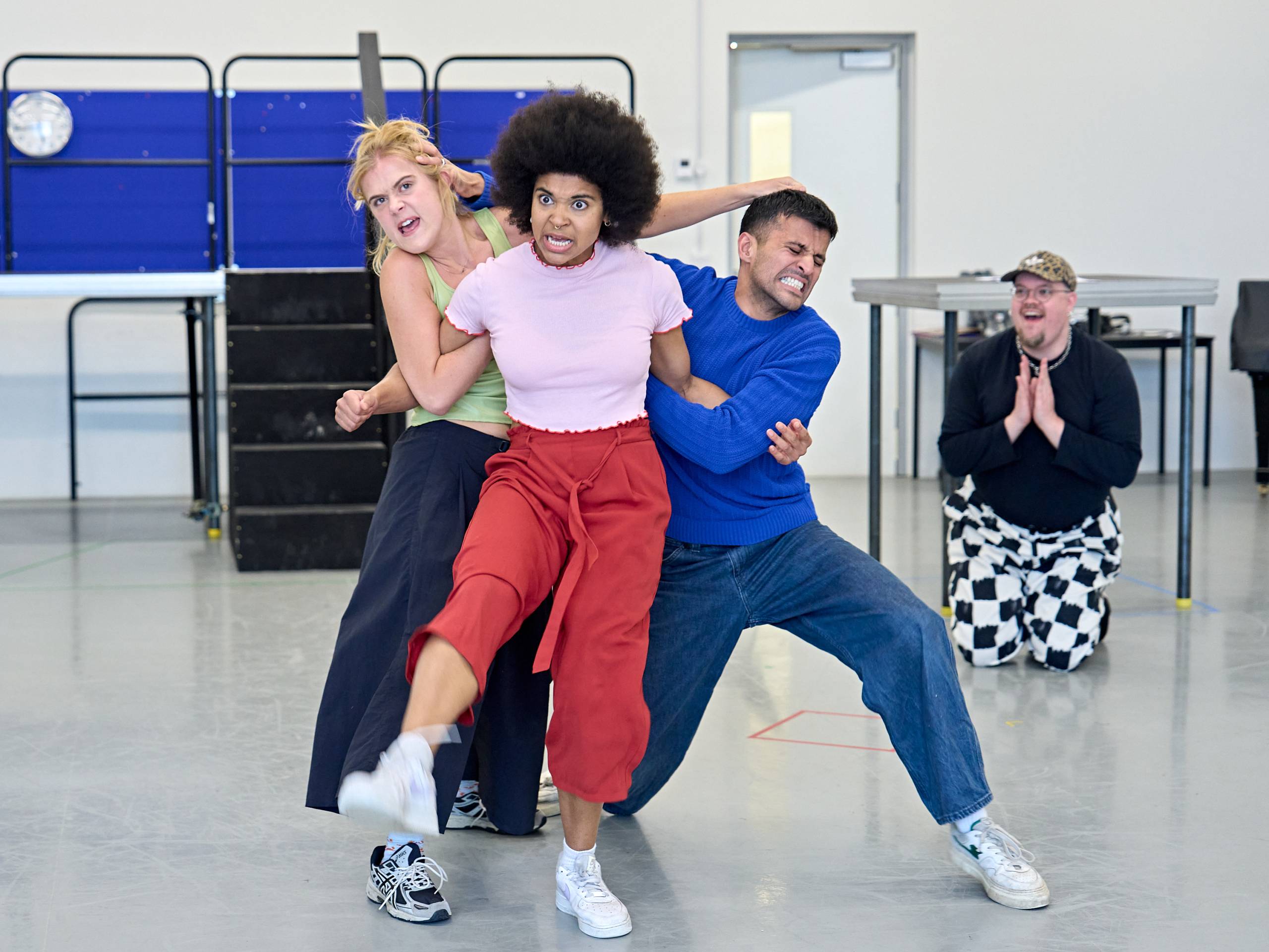 A group of performers on a stage engage in an energetic rehearsal. One individual in the centre is held by two others and appears animated, with dynamic body language. Another person in patterned pants kneels in the background, hands clasped, observing the scene with enthusiasm.