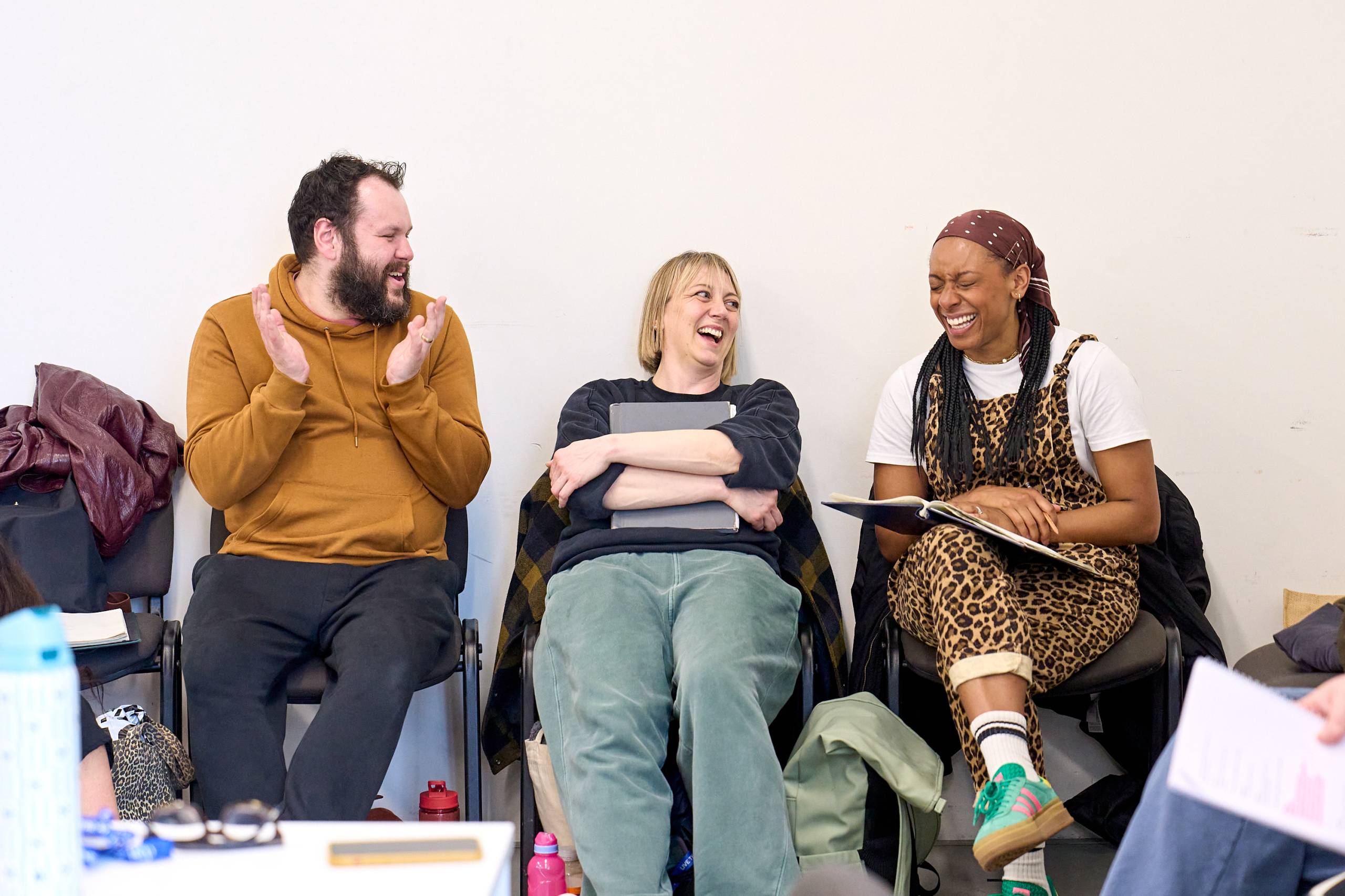A group of three people are sitting on chairs and laughing together in a casual setting. They appear to be engaged in a conversation or participating in an activity. One person is clapping in response to something amusing.