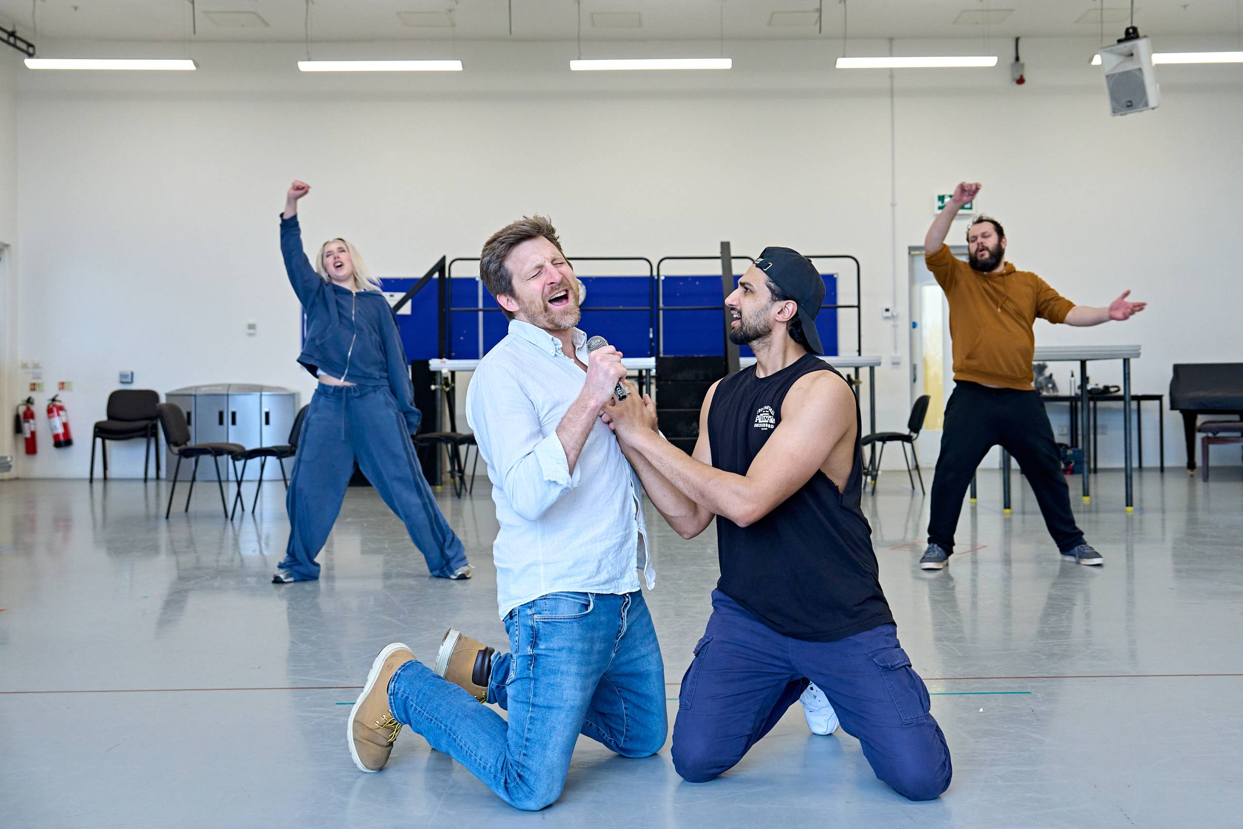 Image of a lively rehearsal scene where four individuals are energetically singing and performing. Two of them kneel in the foreground singing dramatically into a microphone. The atmosphere is animated and joyful.