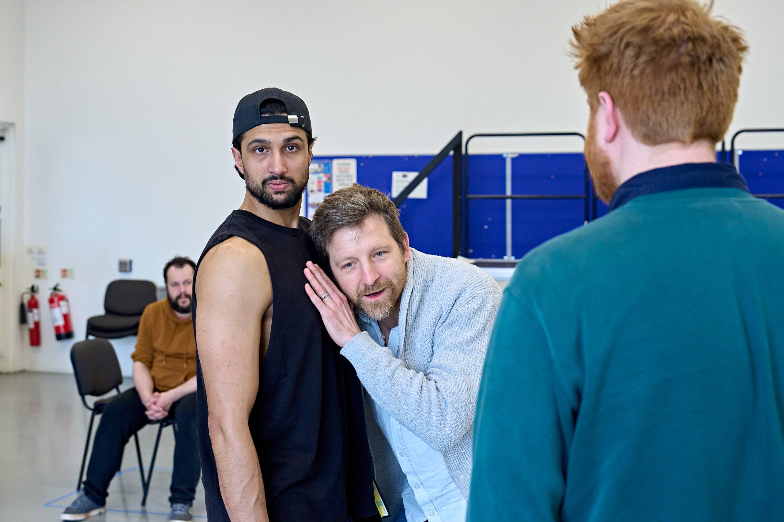 A person wearing a sleeveless shirt is standing staring directly into the camera while another person leans in to their chest, appearing to listen to their heart. A third person with a beard stands nearby, observing.