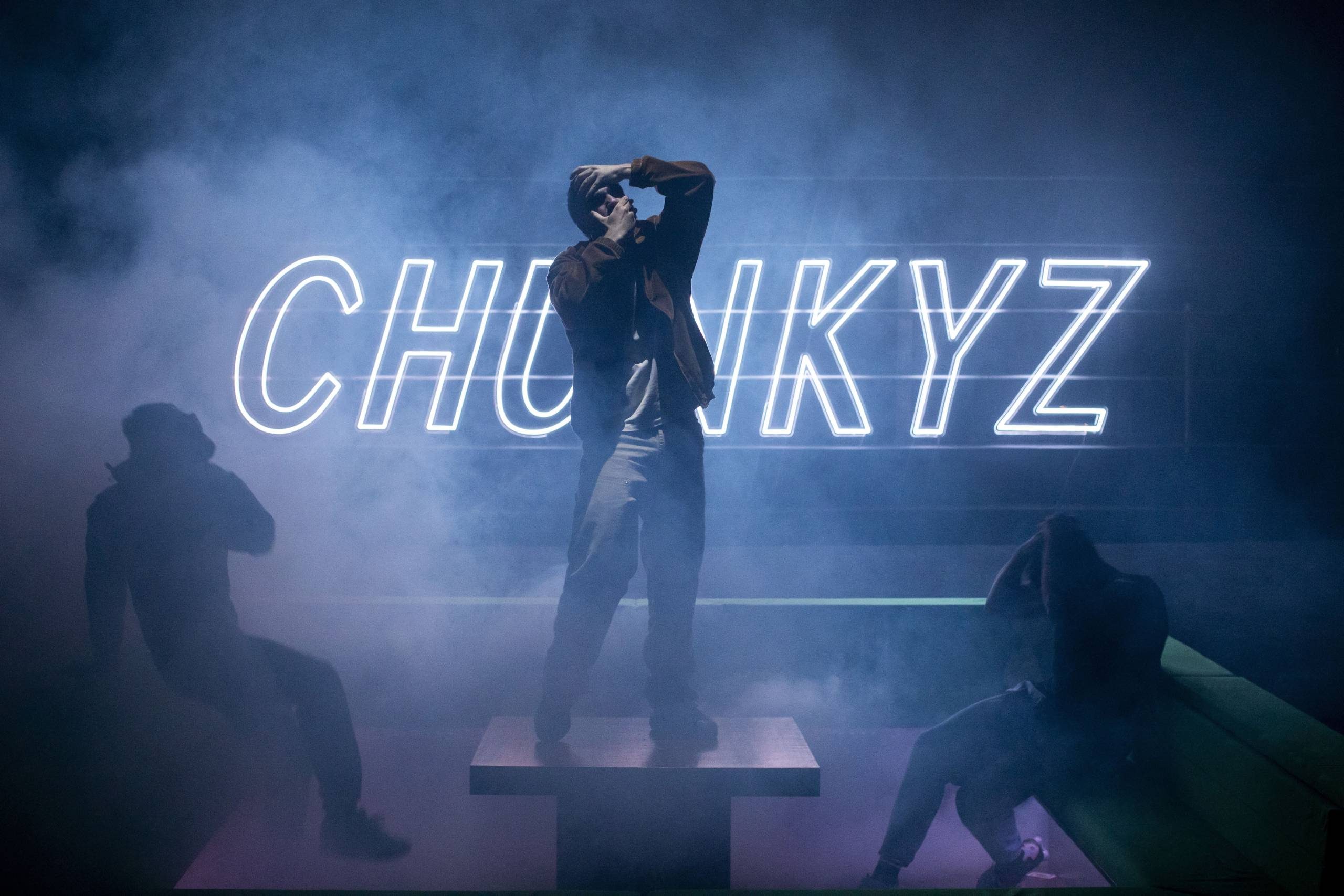 Three people pose in a foggy room with a glowing neon sign reading 