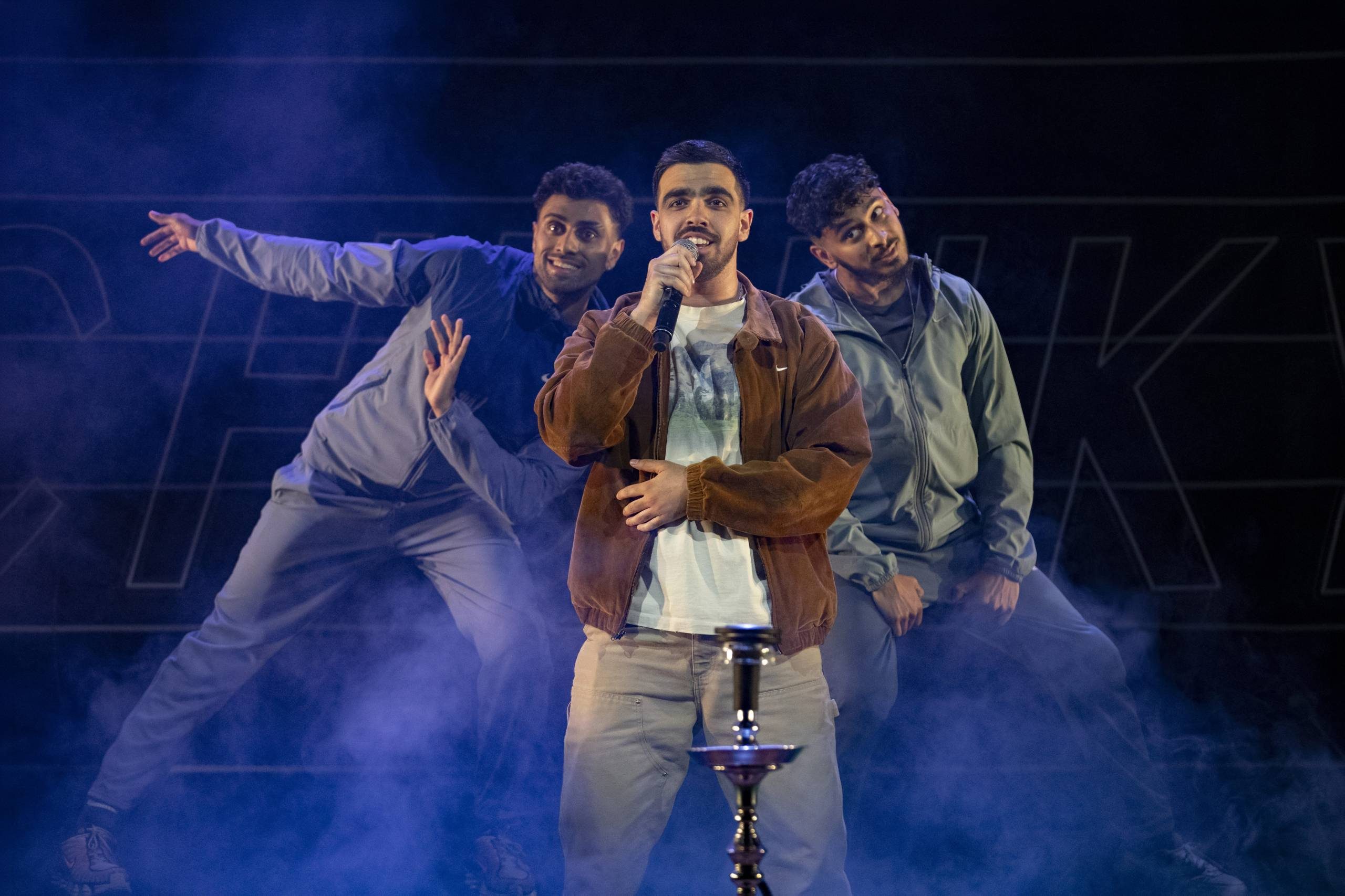Three men performing on stage with one singing into a microphone and two dancing amidst blue smoke effects.
