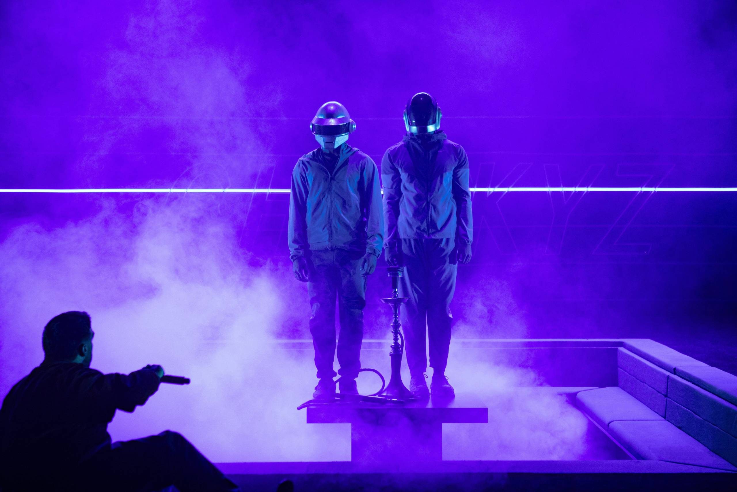 Two figures in robotic helmets stand on a platform amidst purple smoke and lighting, with a seated person in silhouette holding a microphone in the foreground.