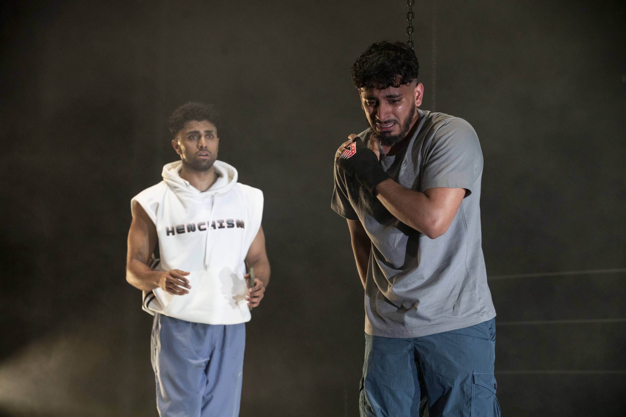 Two men on stage, one in a white hoodie and the other in a gray shirt, with dramatic lighting and a sombre atmosphere.
