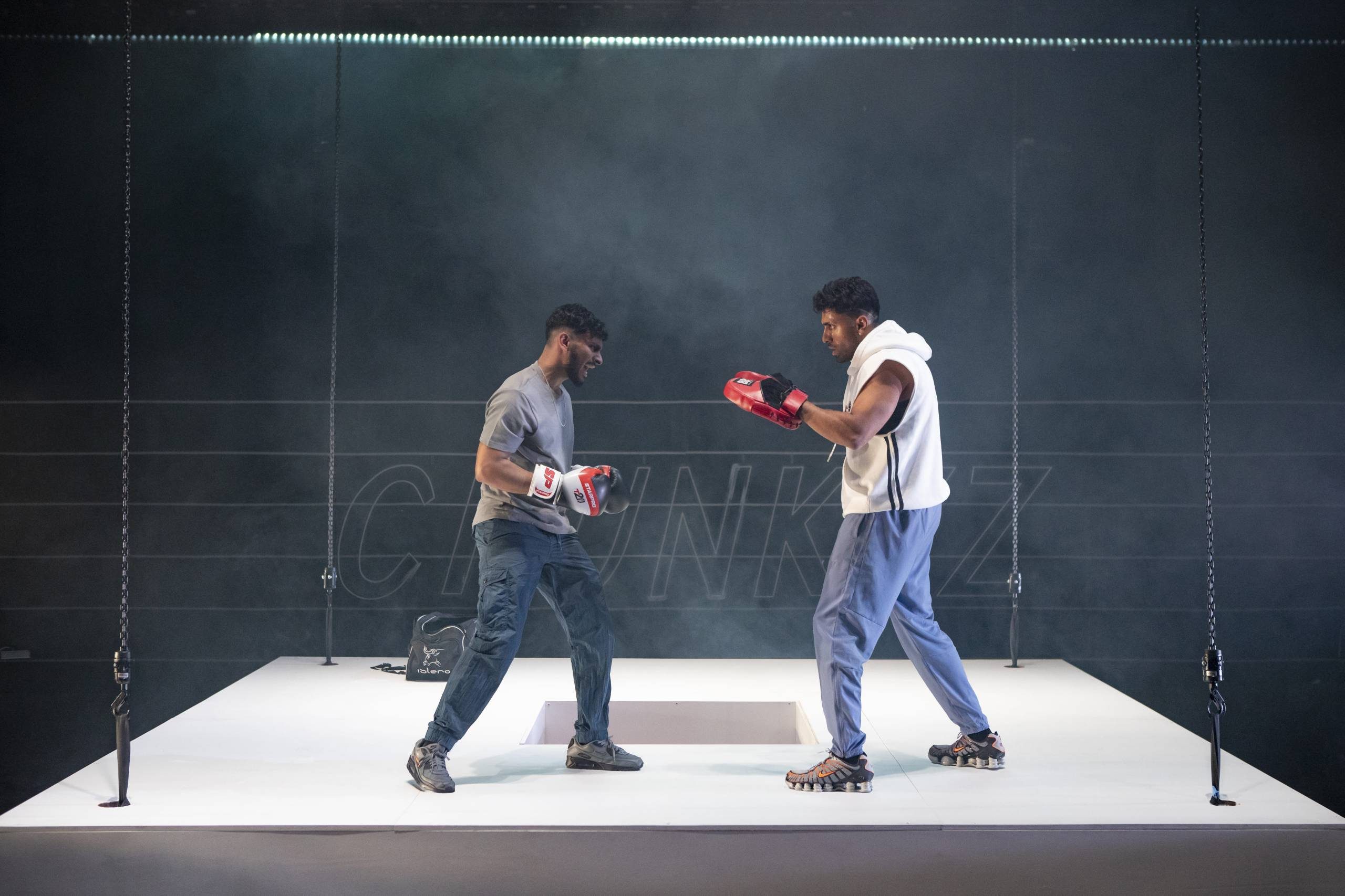 Two men wearing boxing gloves spar on a suspended white platform in a dim, smoky environment.