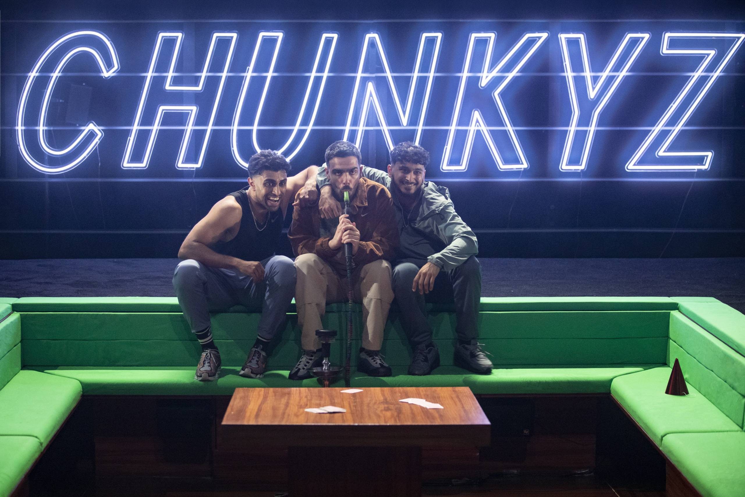 Three men sit on a green sofa in front of a neon 