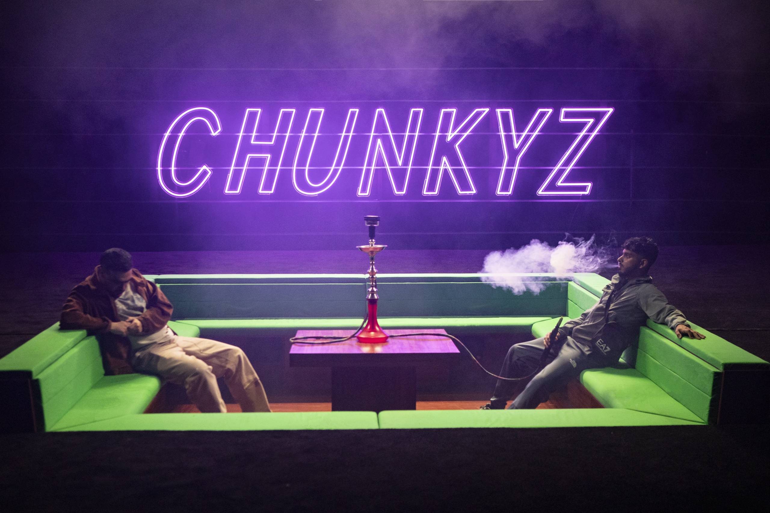 Two men sit on a green couch around a hookah with smoke billowing out under a neon 
