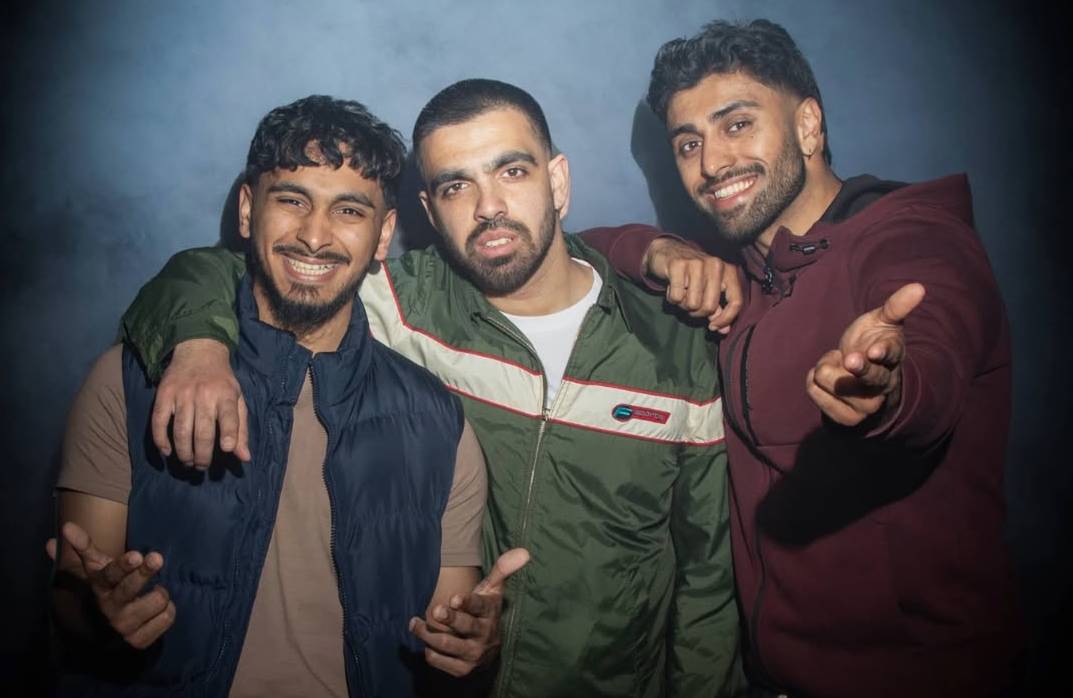 Three young men are standing together against a dark, smoky background. Two are wearing casual jackets while the third has a hoodie. They appear cheerful, with one extending a hand towards the camera.