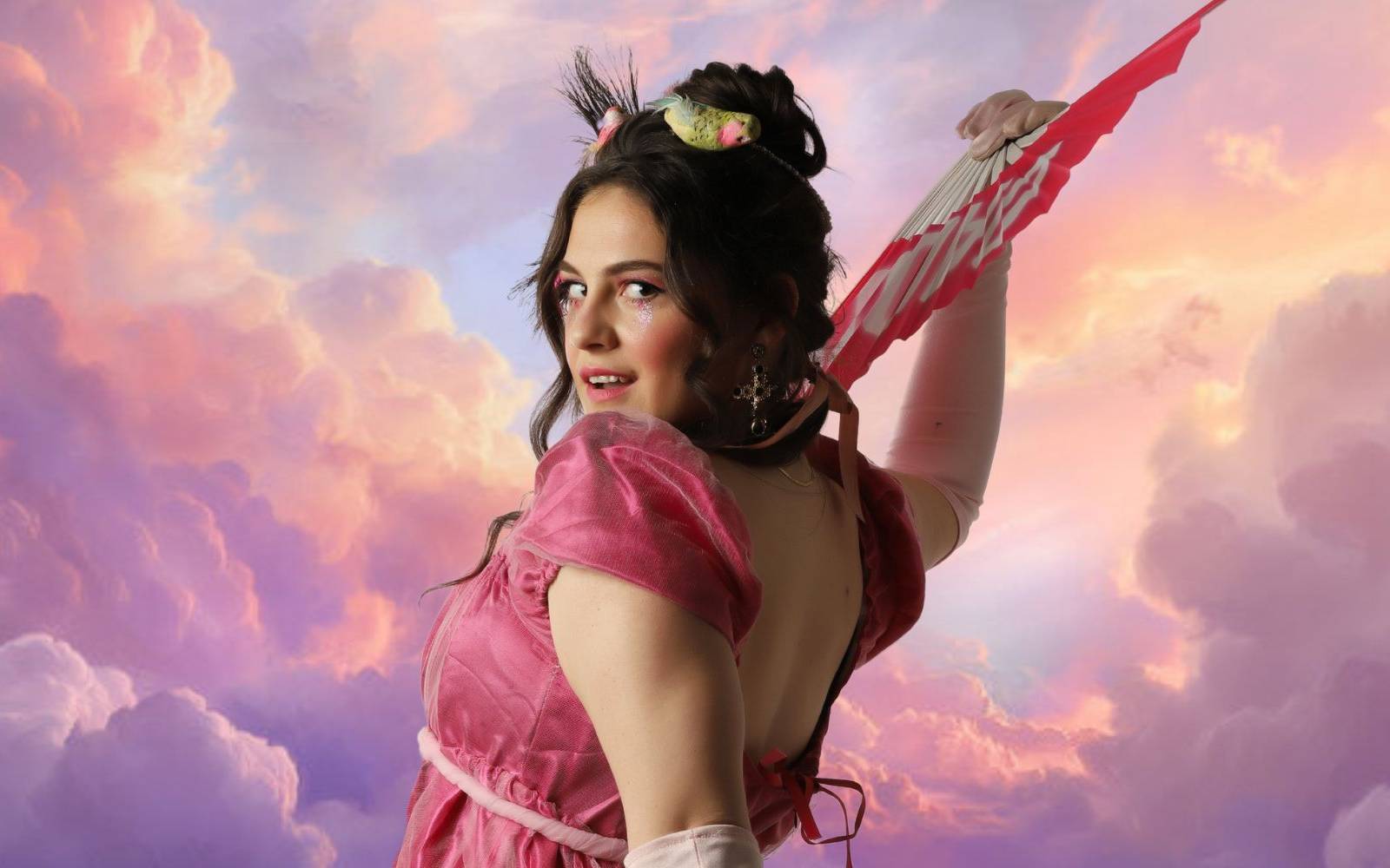 A woman in a pink, vintage-style dress holds a red fan against a vibrant sunset sky with pastel clouds.