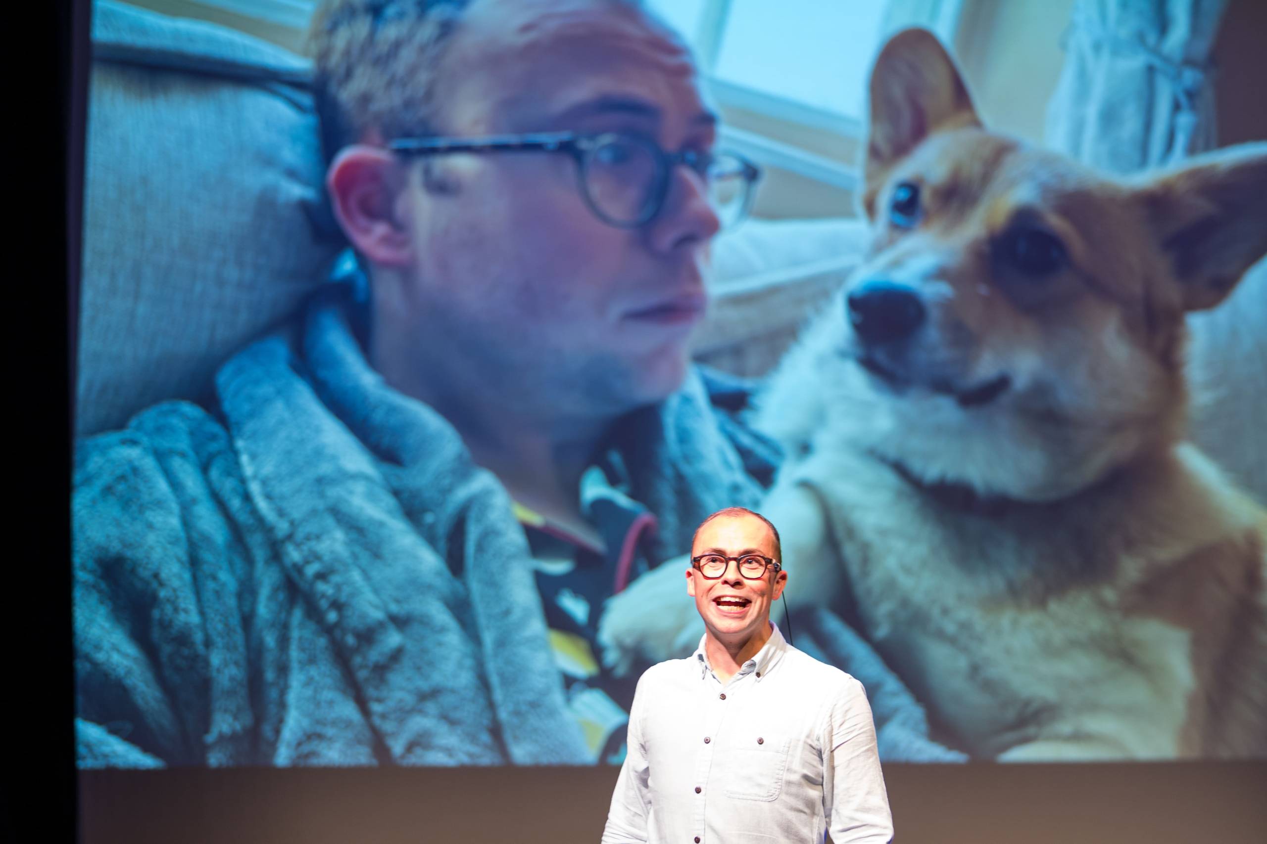 Joe Tracini wears a black shirt and jeans, behind him a screen show a photograph of Joe cuddling a dog.