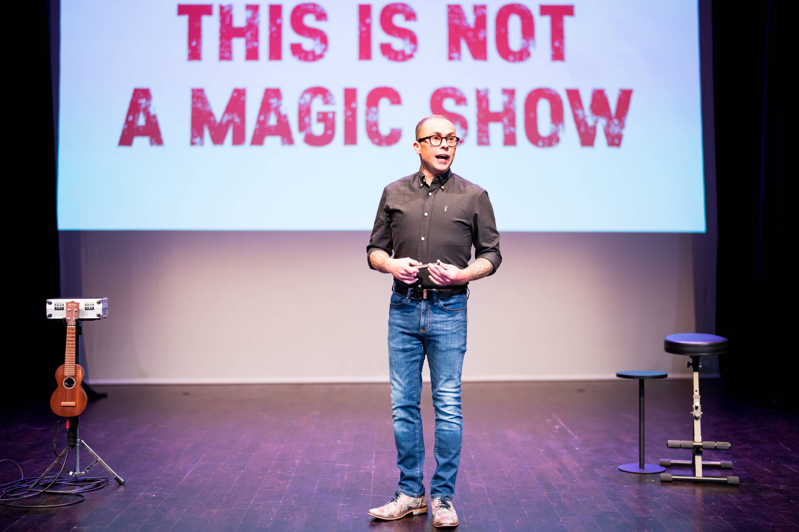 Joe Tracini wears a black shirt and jeans, behind him a screen reads THIS IS NOT A MAGIC SHOW.