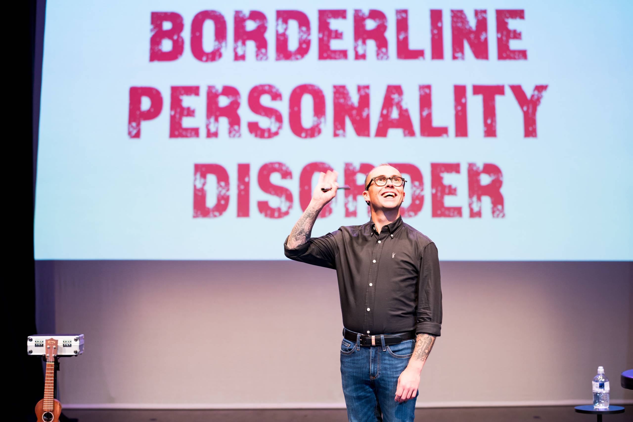 Joe Tracini wears a black shirt and jeans, behind him a screen reads BORDERLINE PERSONALITY DISORDER.