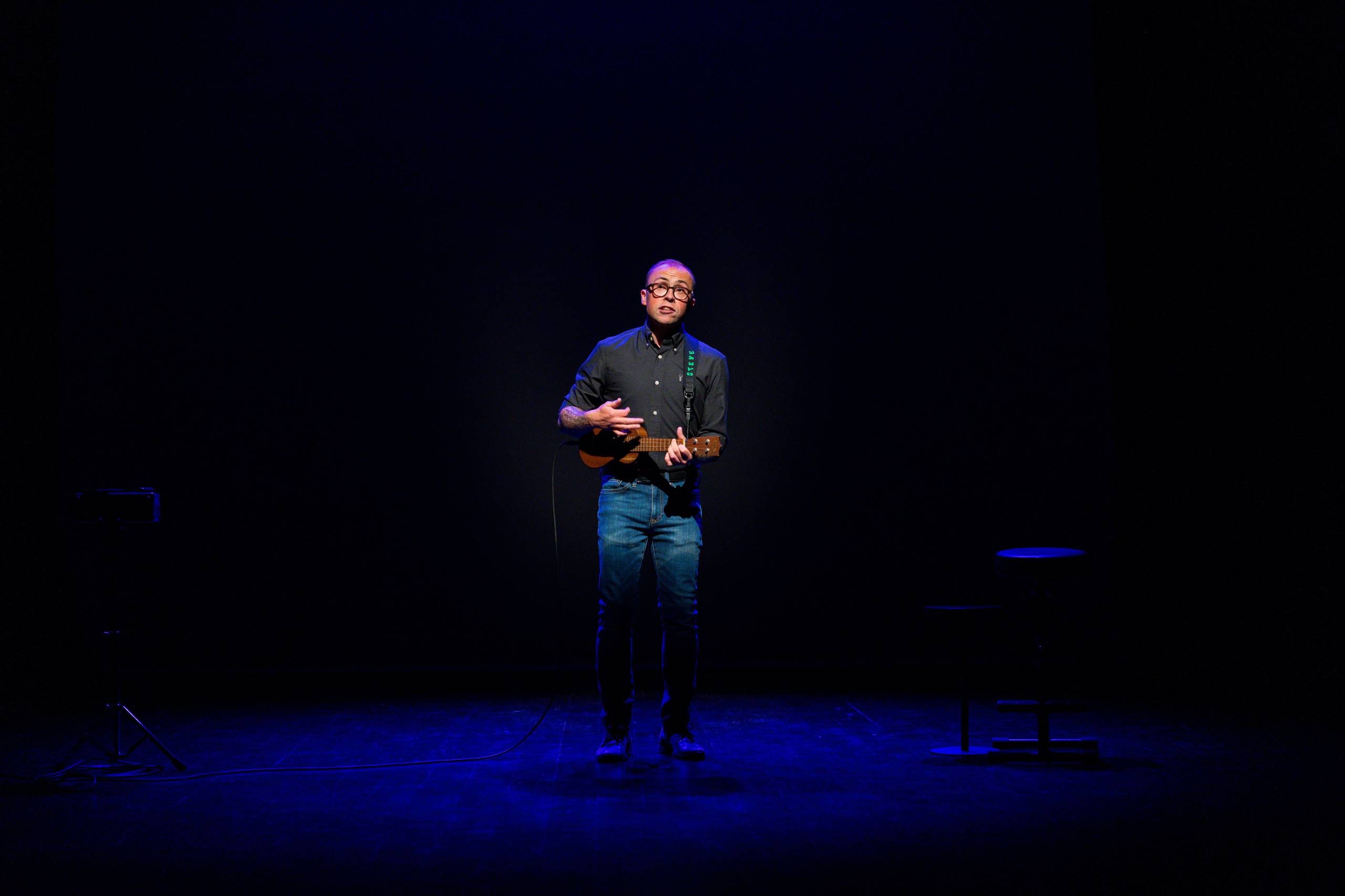 Joe Tracini wears a black shirt and jeans and plays a ukulele in a blue spotlight.