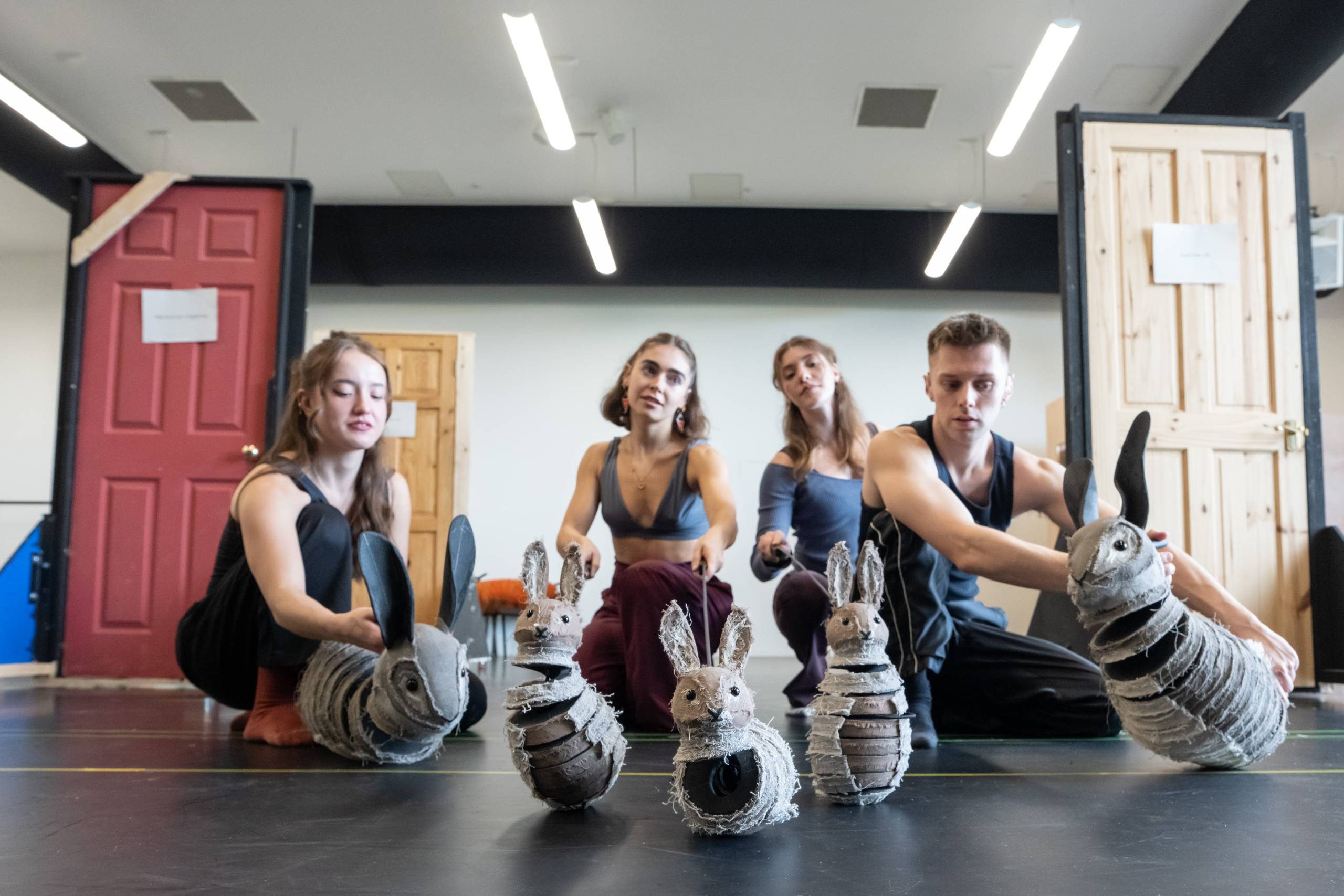 A group of dancers are seated on a studio floor, each holding a rabbit puppet. The room has several wooden doors and bright overhead lighting.