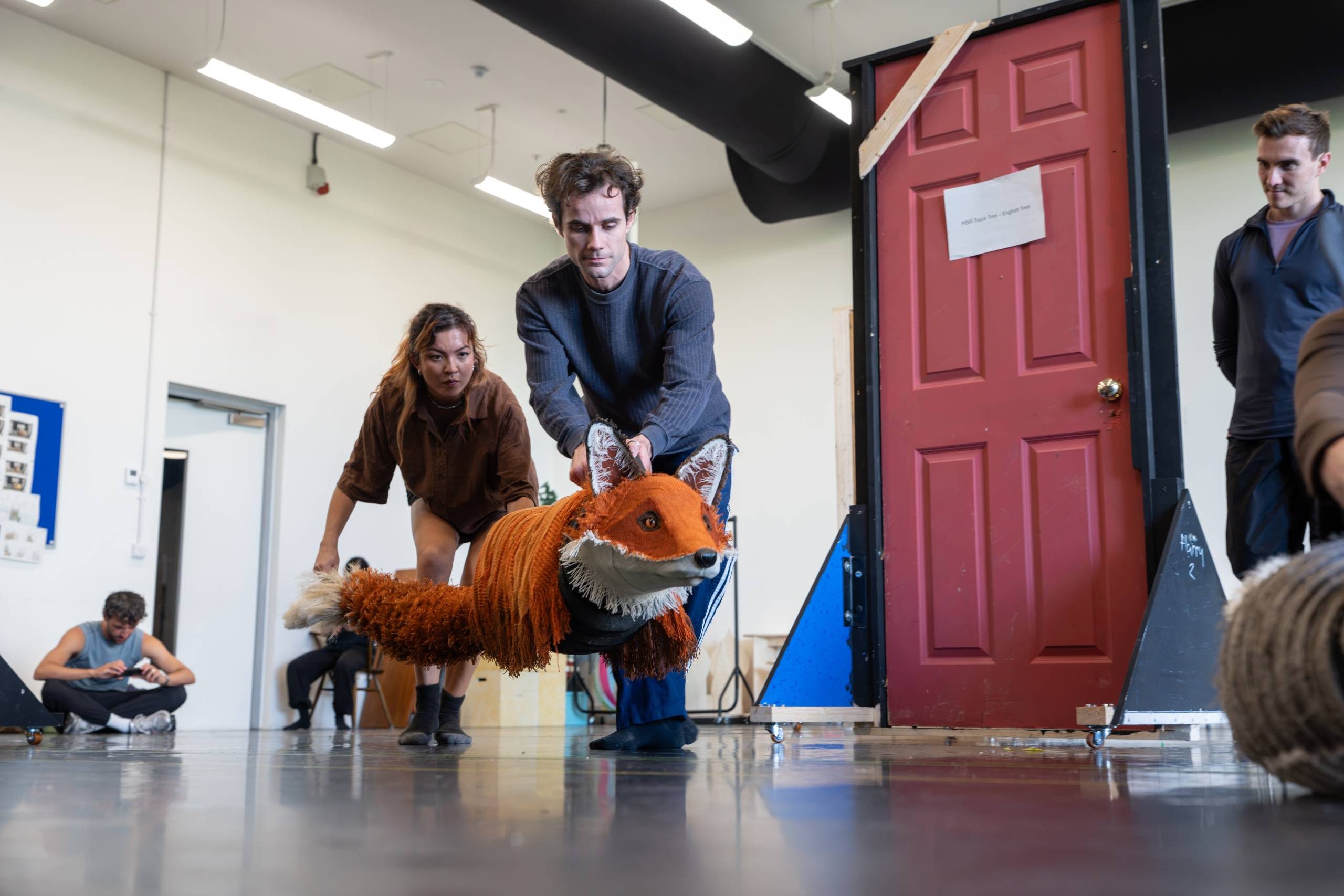Two people are manipulating a large puppet resembling a fox in a rehearsal space. A red door set piece is in the background.