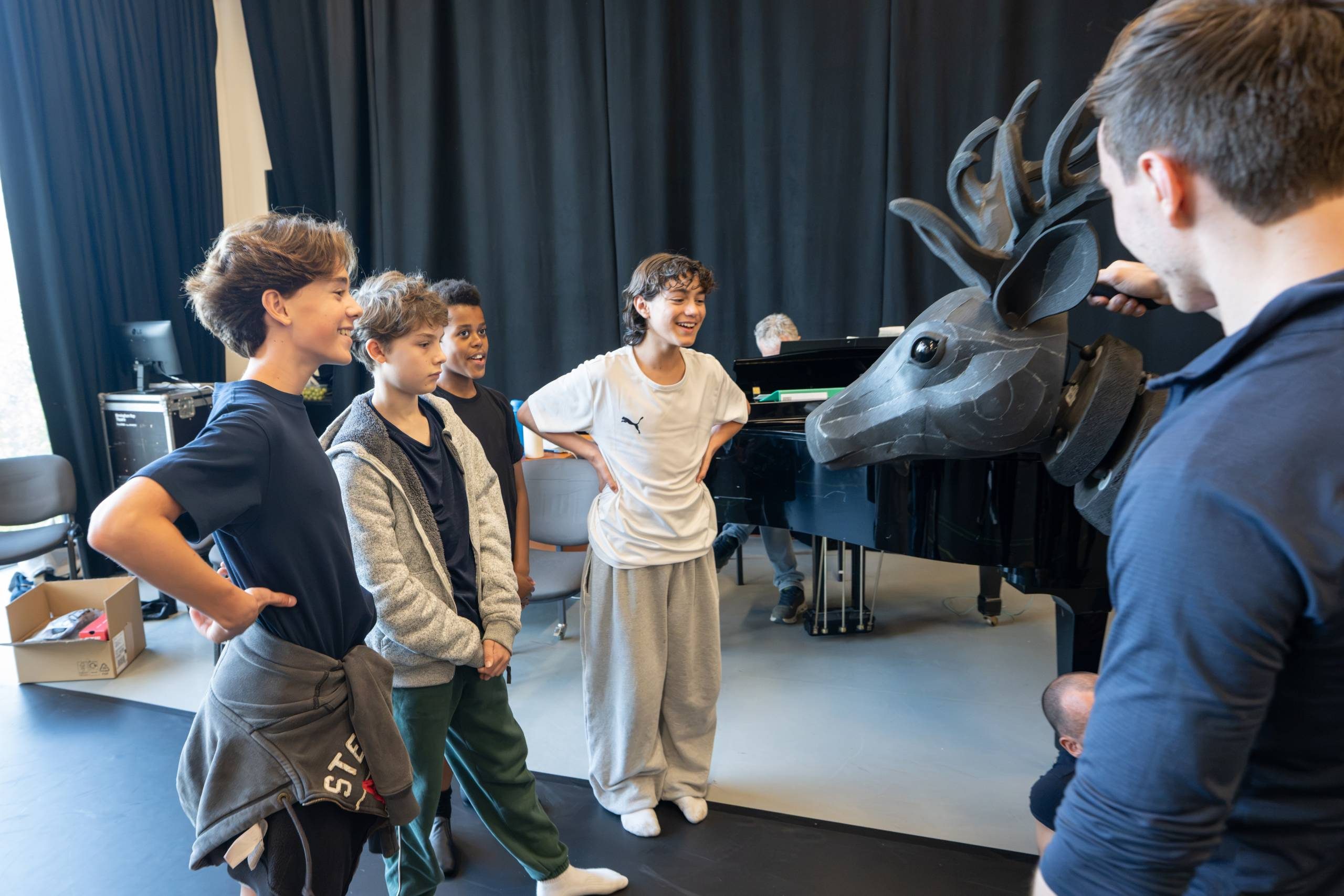 A group of child actors stand around a large, stylized deer puppet. Behind them is a piano and chairs, and one person appears to be manoeuvring the deer's head.