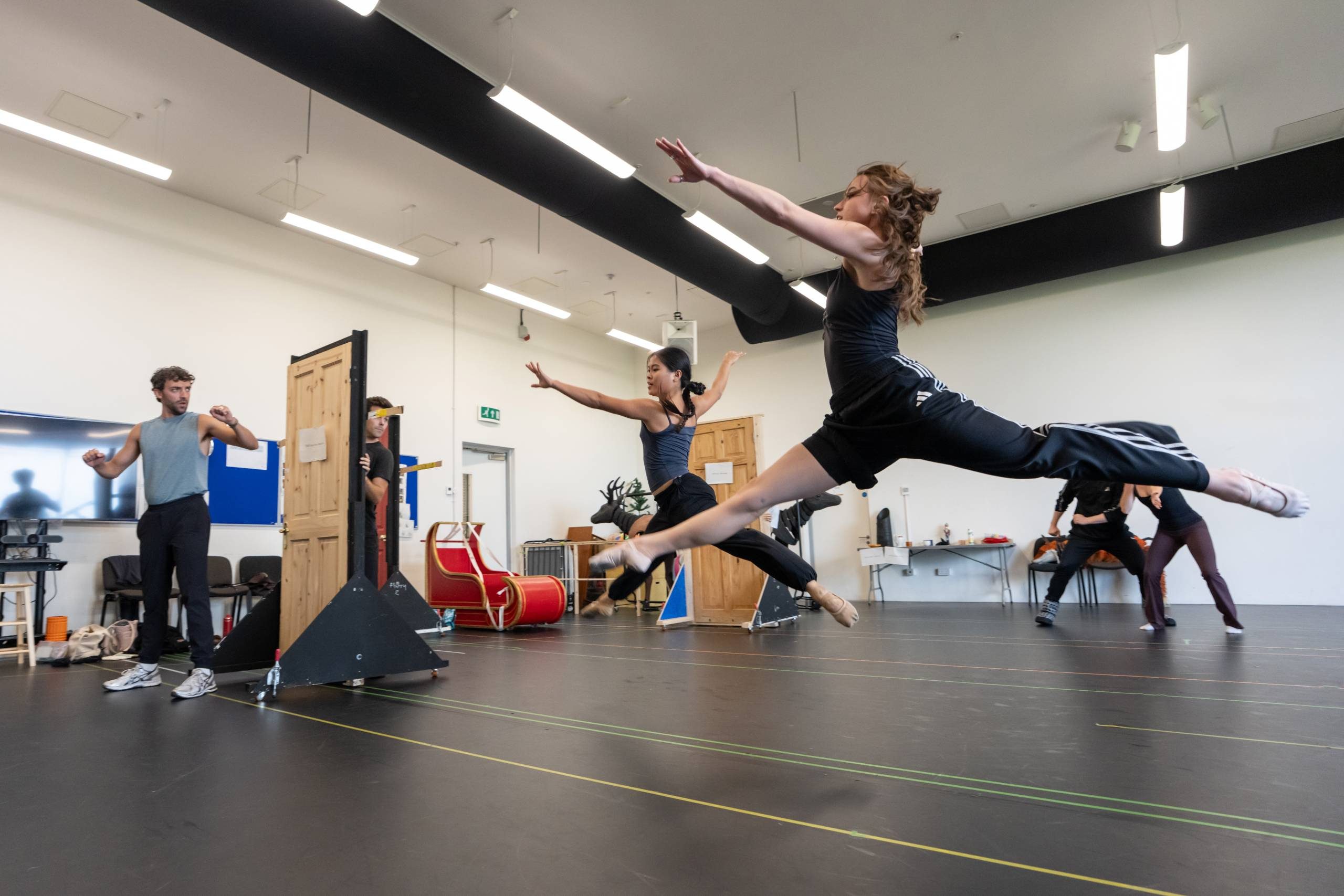 In a spacious rehearsal room, two people are mid-air performing grand jetés, while others observe. The setting includes mirrors, a door prop, and various props scattered around.