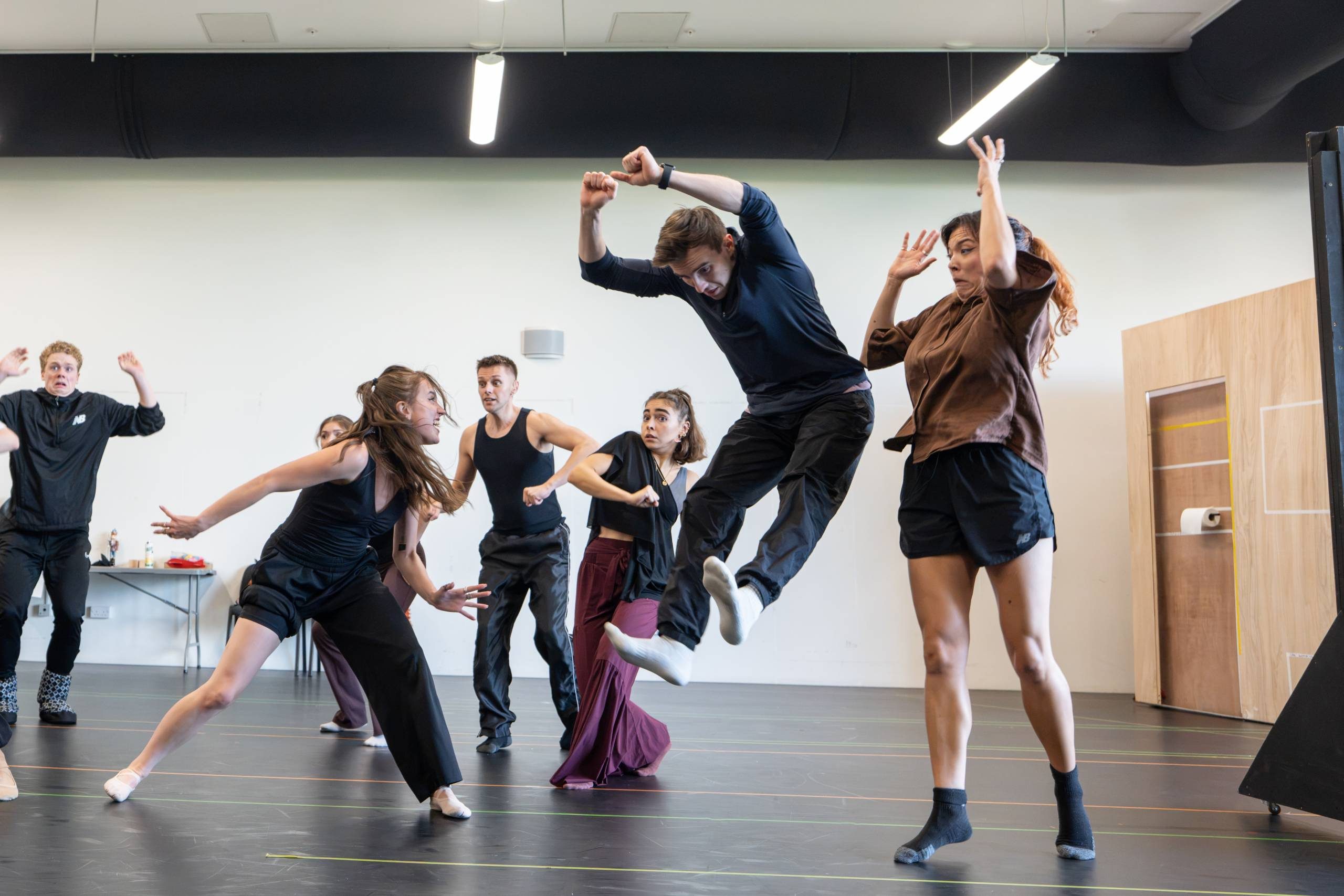 A group of energetic dancers performing in a rehearsal space, captured in mid-motion. Their expressions show that one of the dancers is pretending to menace or scare them, while the others appear frightened and jumping out of the way.