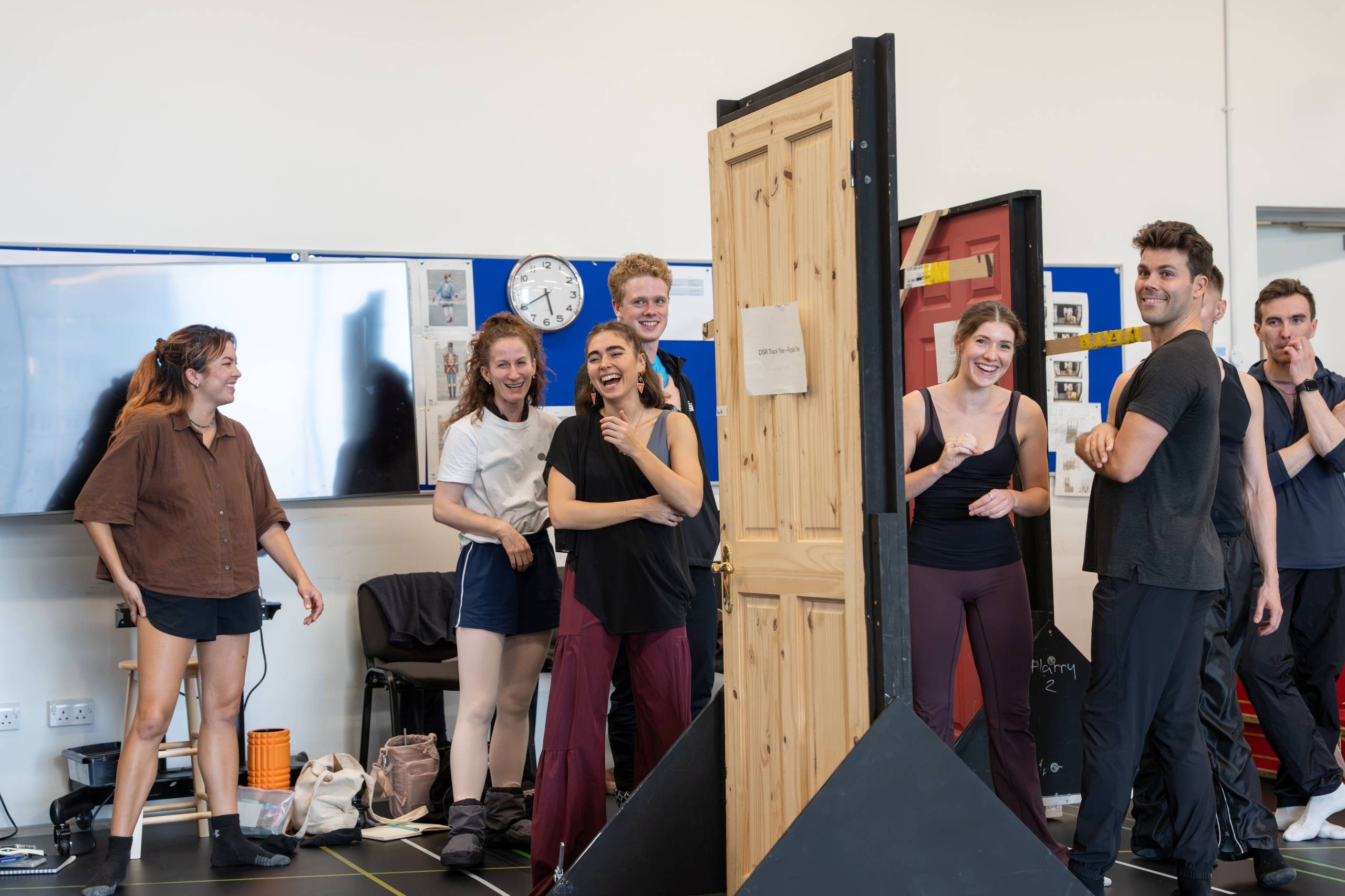 A group of dancers are joyfully standing around a door prop in a rehearsal space. They are casually dressed and appear engaged and cheerful. The background features a wall with a blackboard, whiteboard, clock, and some posted notes.