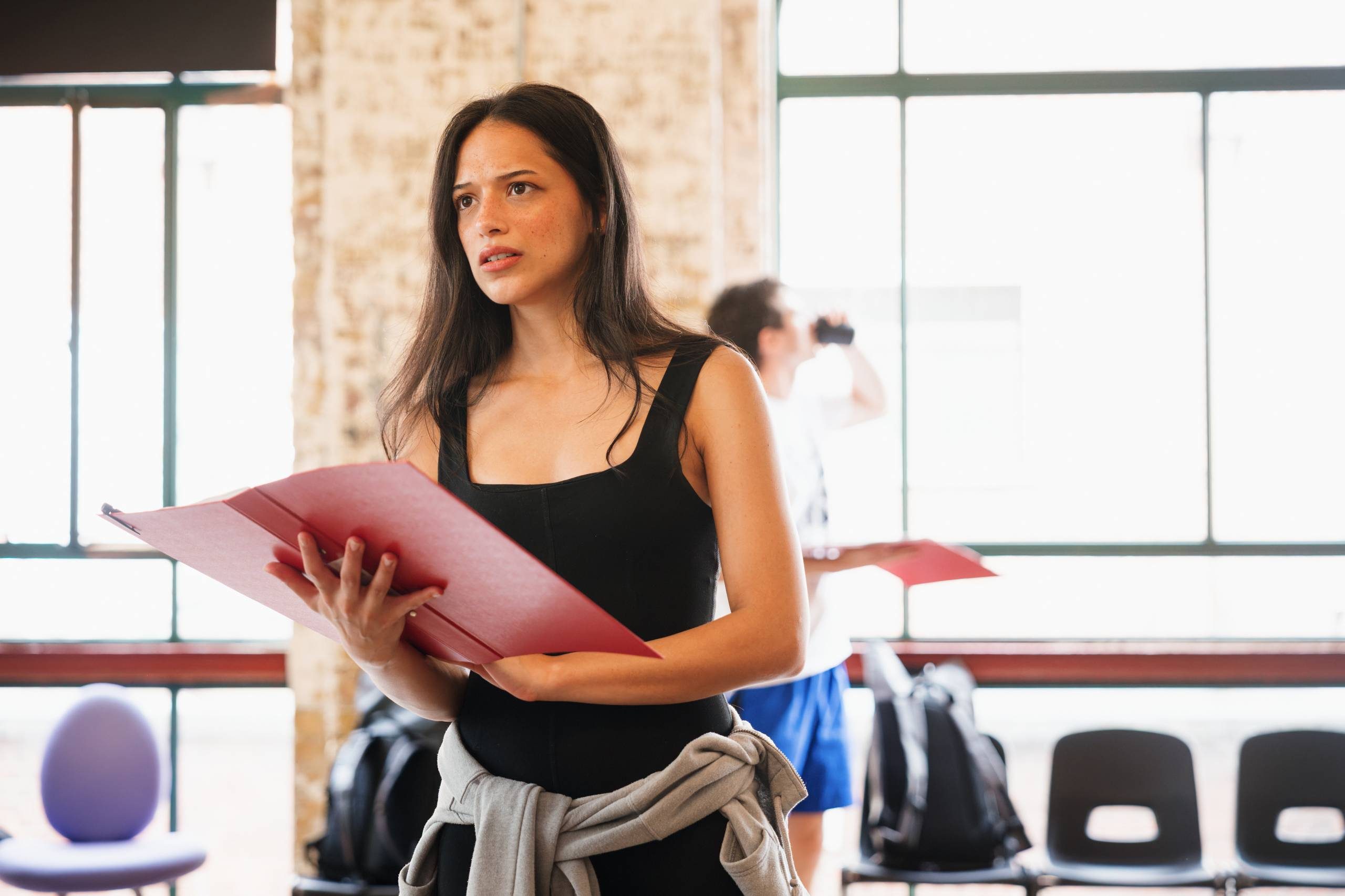 The photo shows a person holding a red folder, standing in a bright room with large windows. Another person is visible in the background.