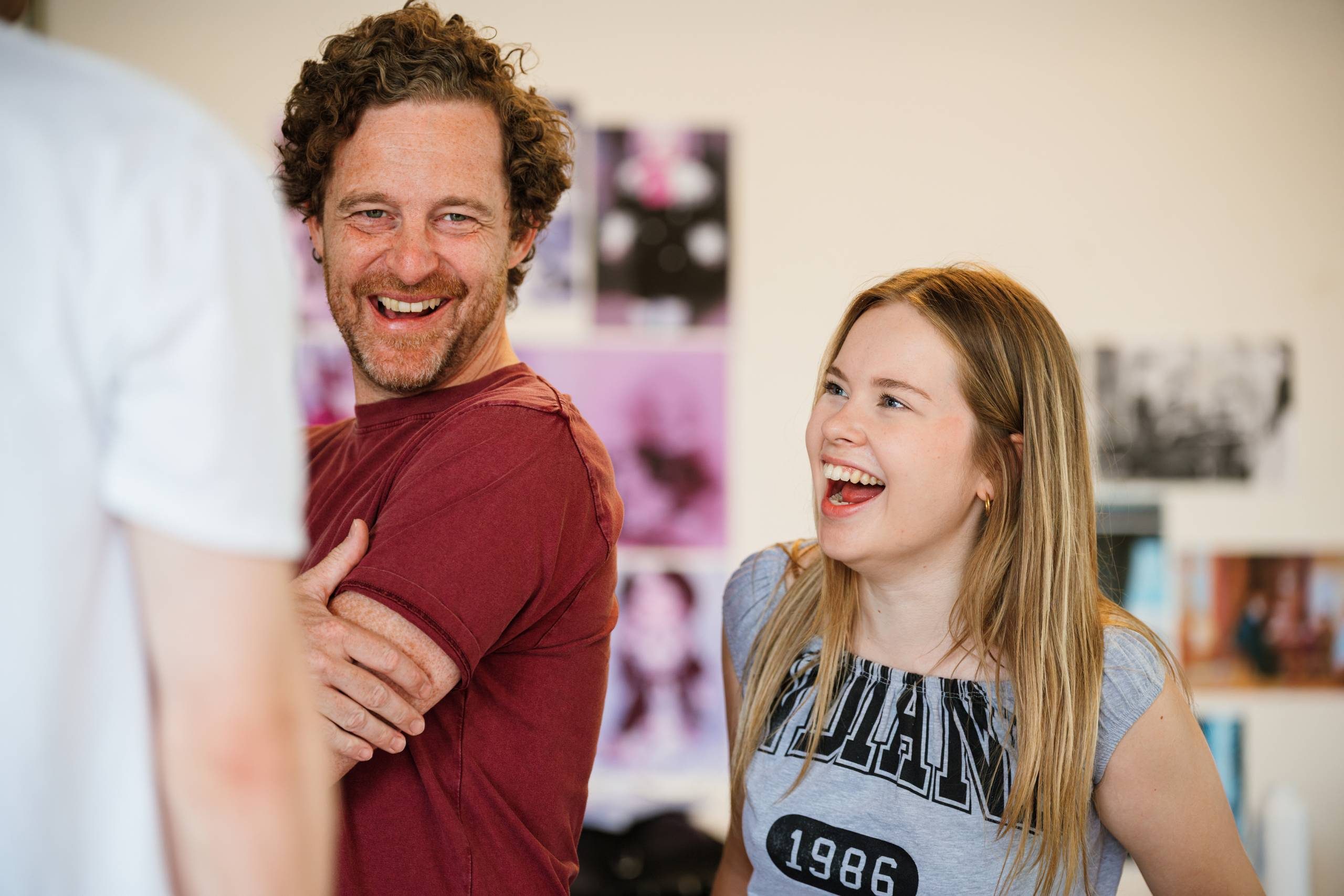 A person with brown curly hair and a person with long blonde hair are smiling in a lively rehearsal room with posters on the wall.