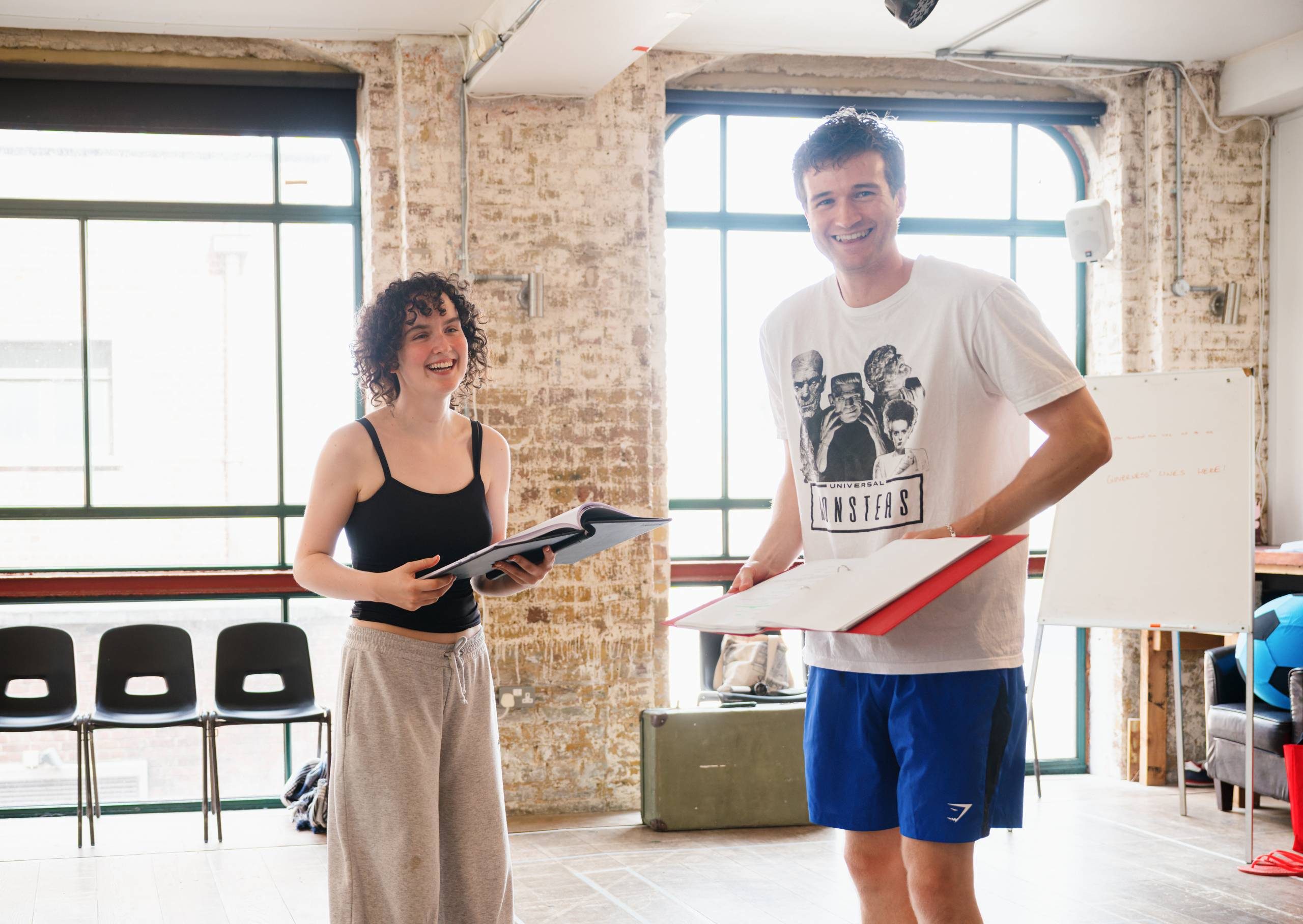 Two people stand in a room with brick walls and large windows, holding scripts. They're smiling and are casually dressed.
