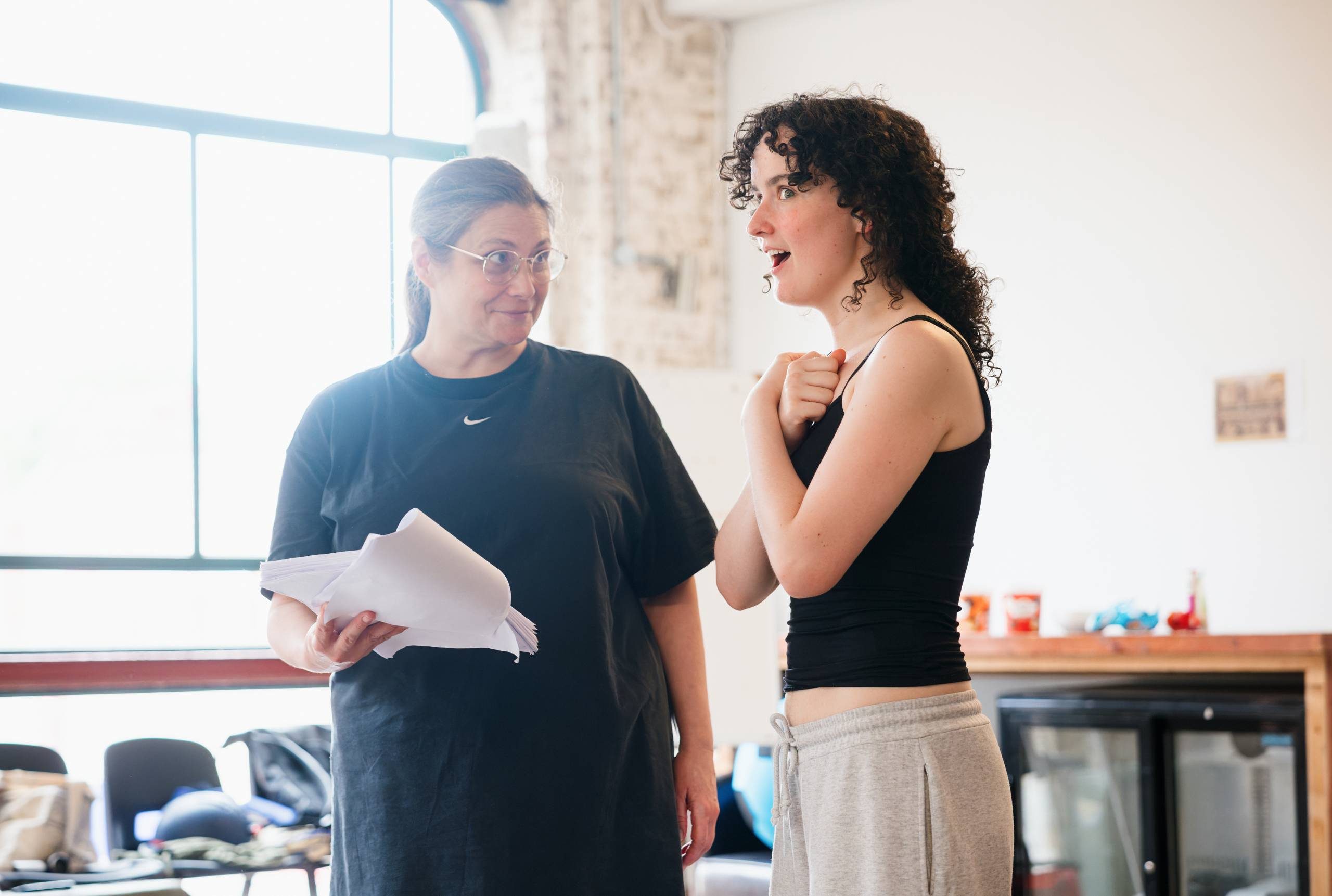 Two people standing in a brightly lit room, possibly a rehearsal space. One person is holding a script and wearing a black Nike shirt. The other is gesturing expressively, wearing a black top and gray pants. A large window and exposed brick wall are in the background.