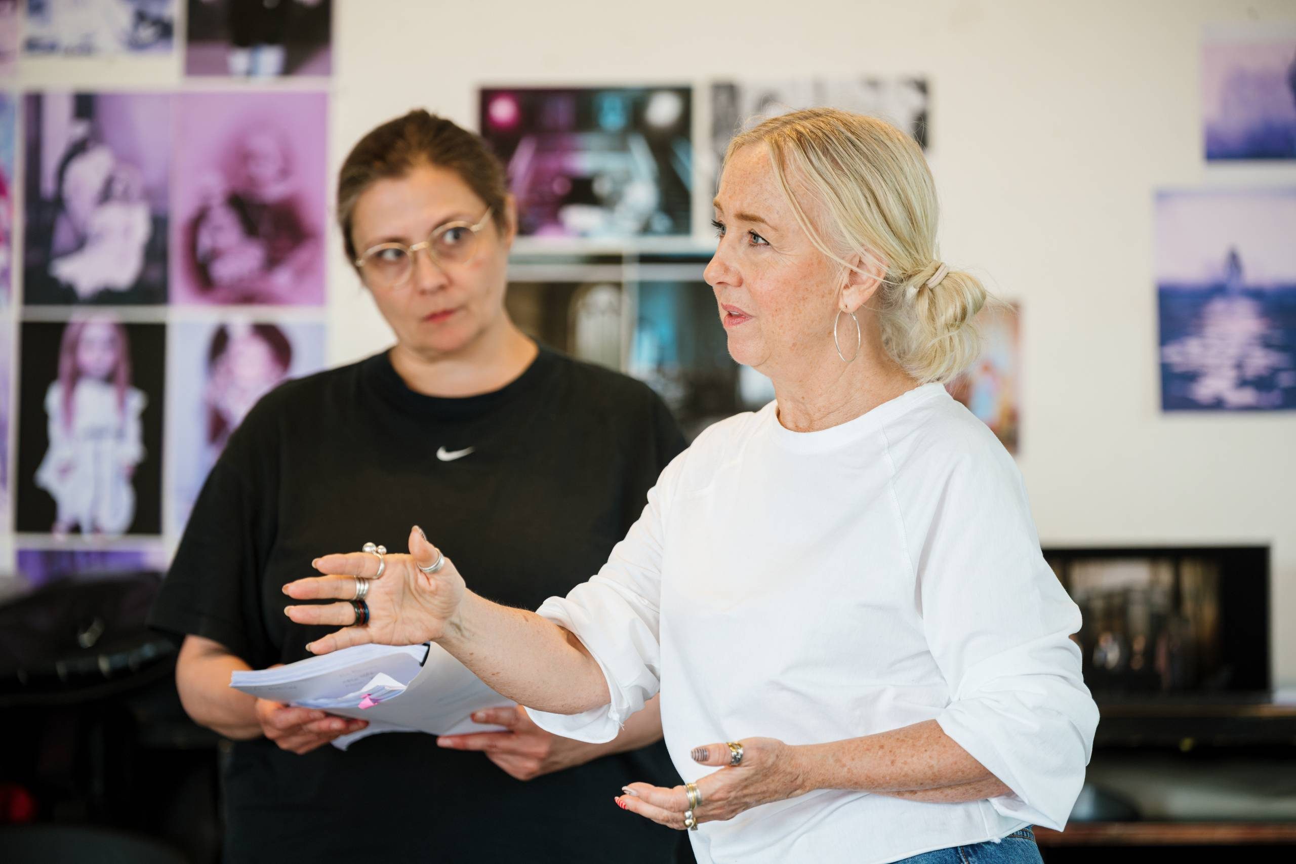 Two people are in a room engaged in conversation. One person is holding a script and listening attentively. There are various photographs on the wall in the background.