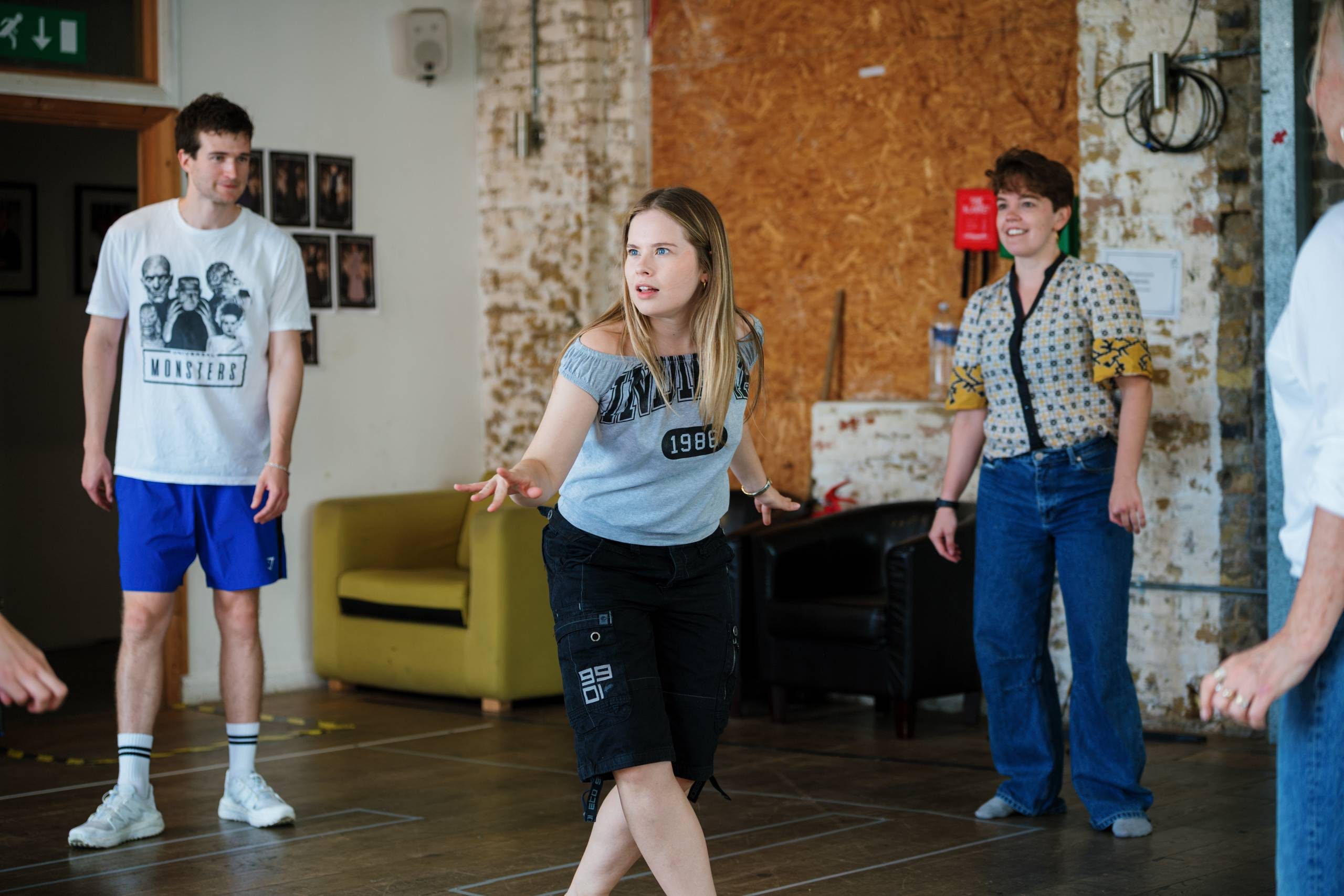 A group of people engaged in a theatre rehearsal, standing in a room with a rustic brick wall. One person is in the foreground, gesturing expressively. Others are watching and standing nearby, casually dressed. The atmosphere is focused and collaborative.