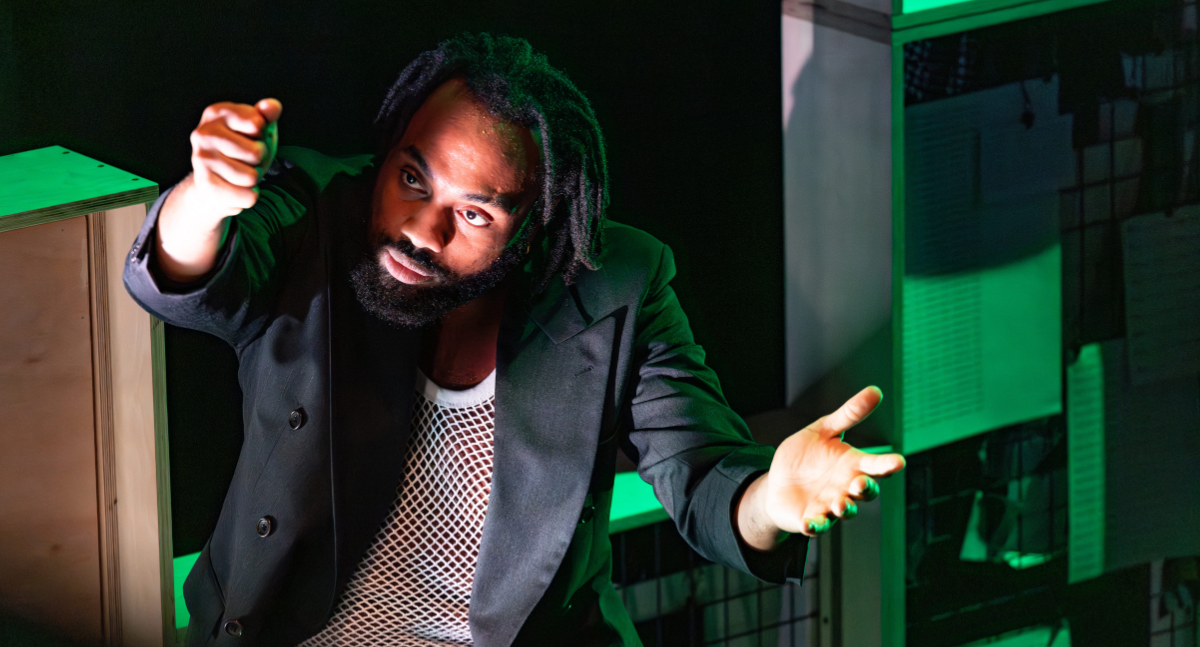 A man with short dreadlocks and a beard gestures expressively, wearing a black blazer and mesh shirt. He's in a dimly lit space with green lighting.