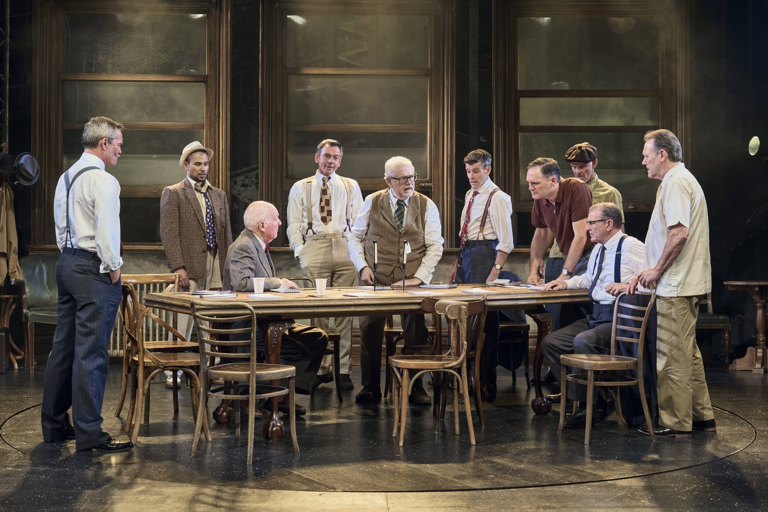 Ten male Jurors are gathered around a table, eight stand while two sit. They are debating the case they must rule on.