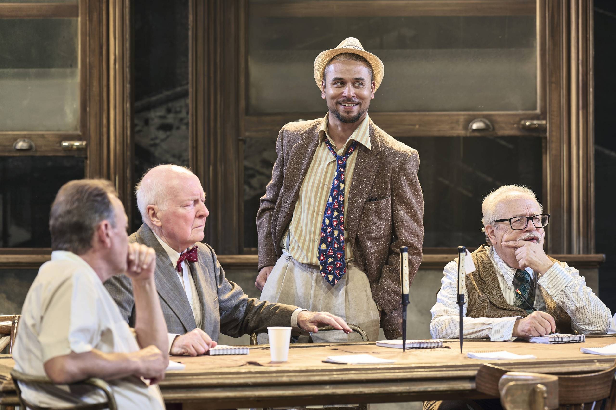 Four male Jurors are gathered around a table, three sit while one stands. The man standing is smiling, with his trilby hat tilted back on his head, his shirt collar is unbuttoned and his tie is loose around his neck. The others look pensive