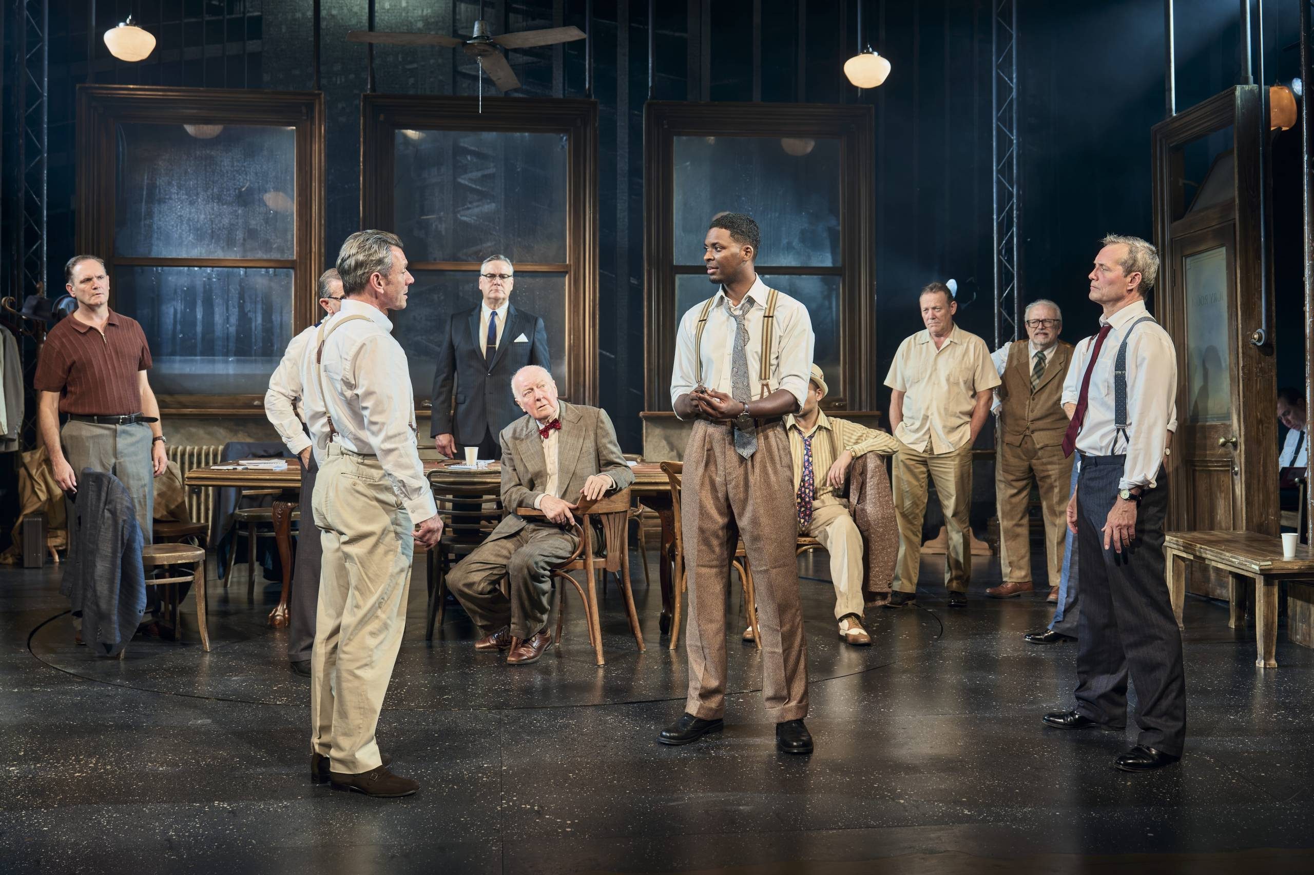 Twelve male Jurors are pictured in a room, ten stand while two sit, they are looking at two man standing who are staring at each other, they look deep in debate. The mood is fraught with tension.