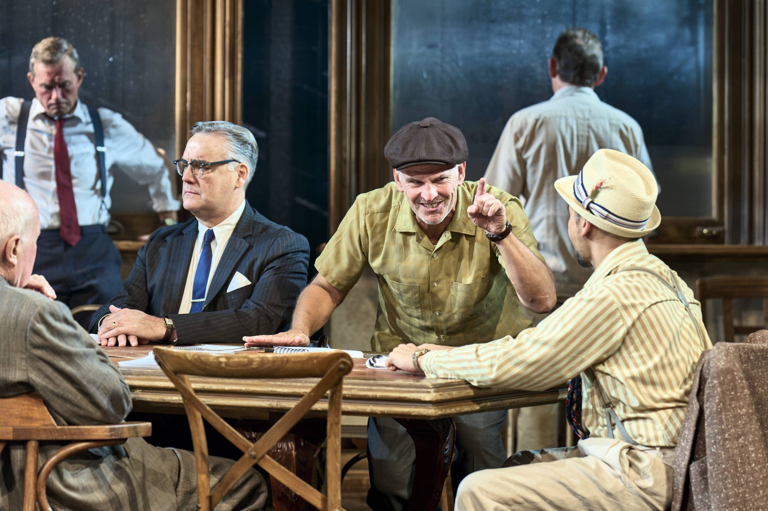 Six male Jurors are pictured in a room, two are standing in the background, one looking out of a window and another staring at the floor. Four Jurors sit around a table, one is animate, his hand is raised and his finger is pointing as he talks to another Juror. the other at the table sit silently.