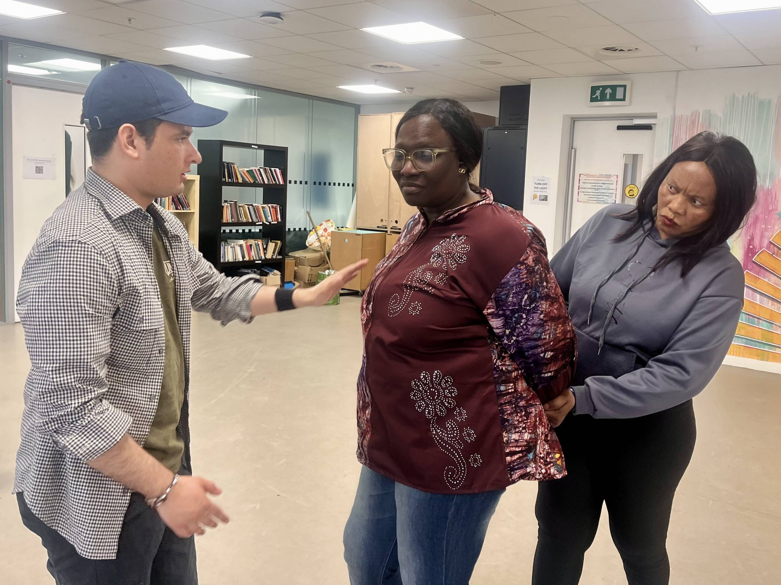 A man wearing a cap gestures as if explaining something to a woman in a patterned blouse, who stands with arms restrained at her side. Another woman stands behind, looking intently, hands on the first woman's back.